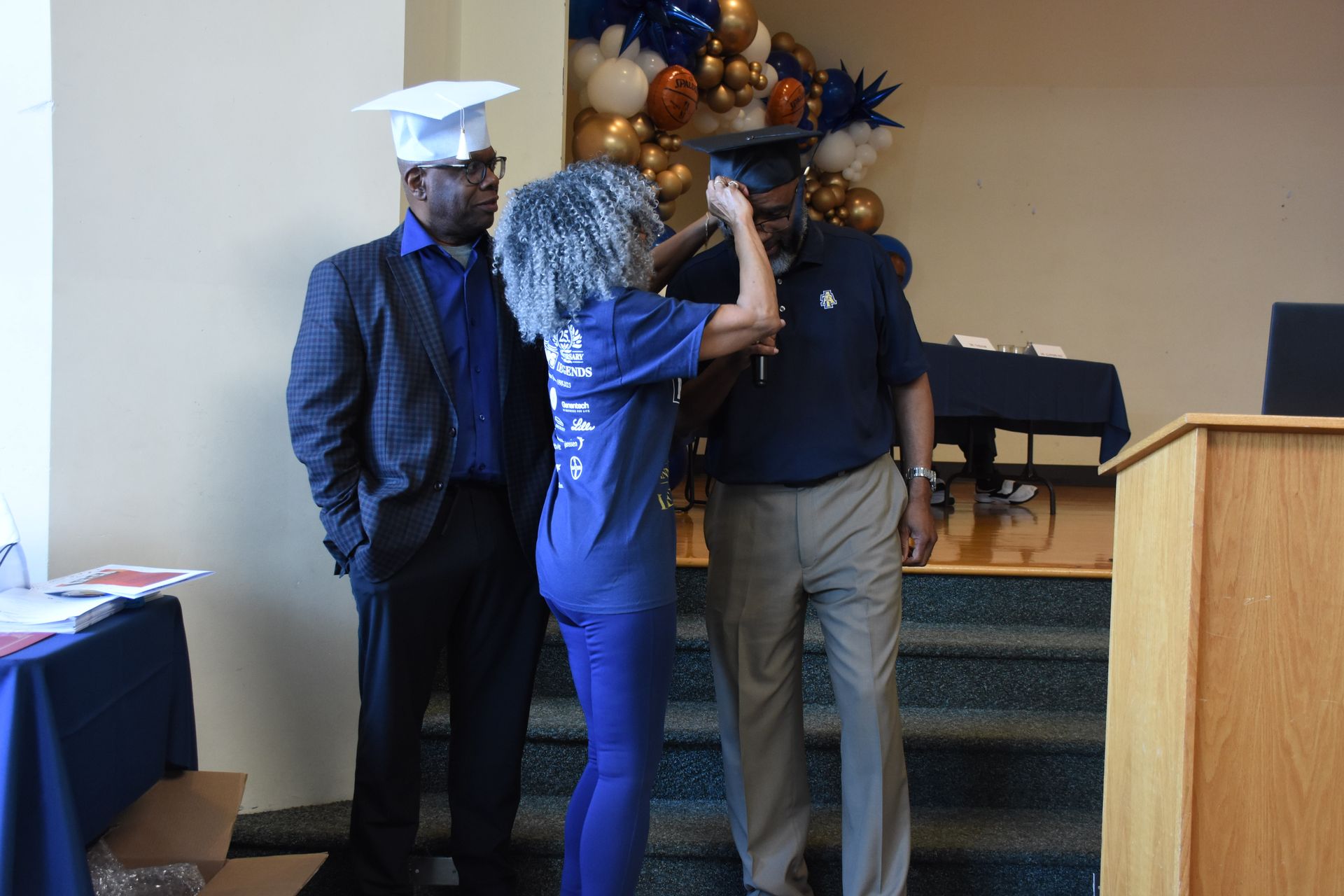 Woman adjusting a graduation cap on a man, another man stands beside them, a blue and gold balloon arch is in the background.