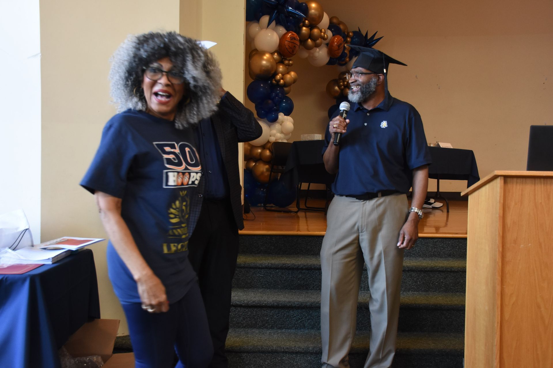 Woman laughing with gray hair and man wearing a cap, standing near podium with balloons in background.