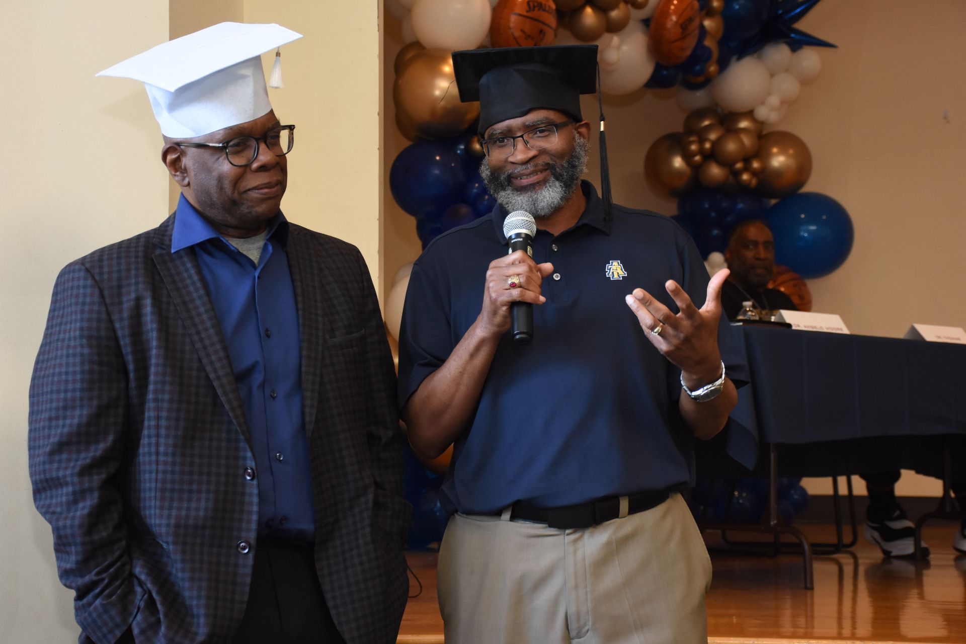 Two men in graduation caps at a podium, one speaking with a microphone, balloons in the background.