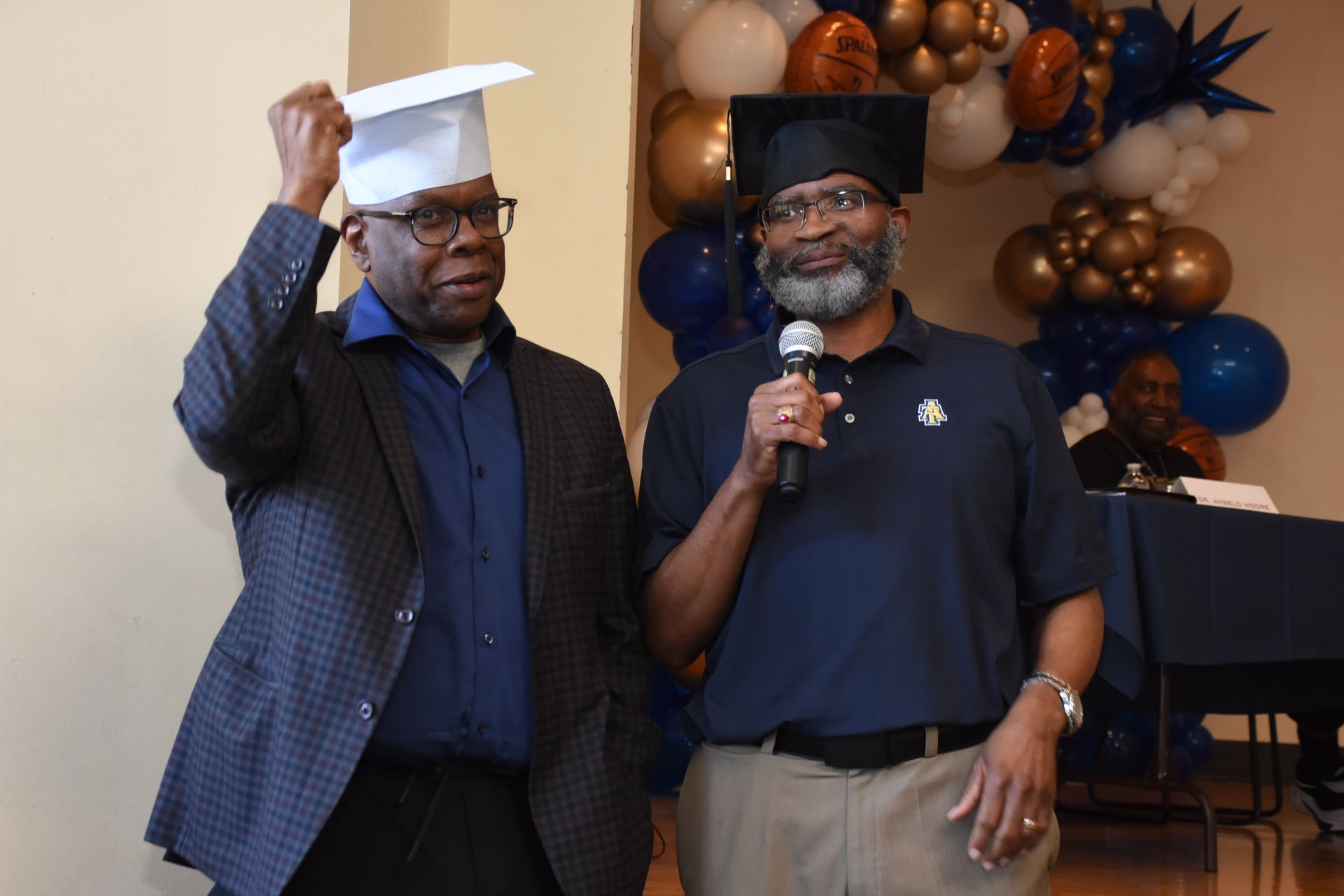 Two men in graduation hats, one holding a microphone, speaking at an event with balloons.