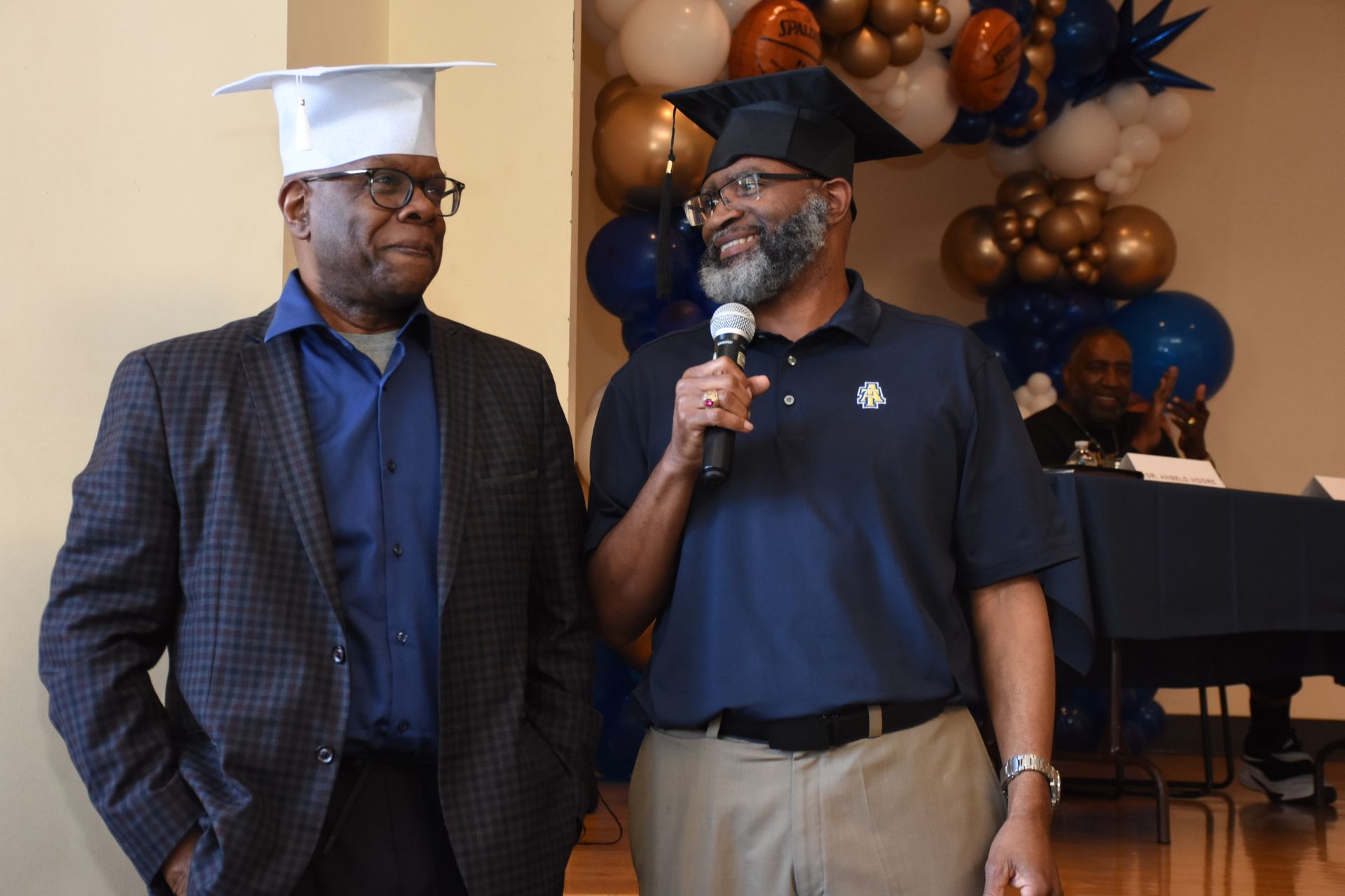 Two men wearing graduation caps, one speaking into a microphone. Balloons in background.