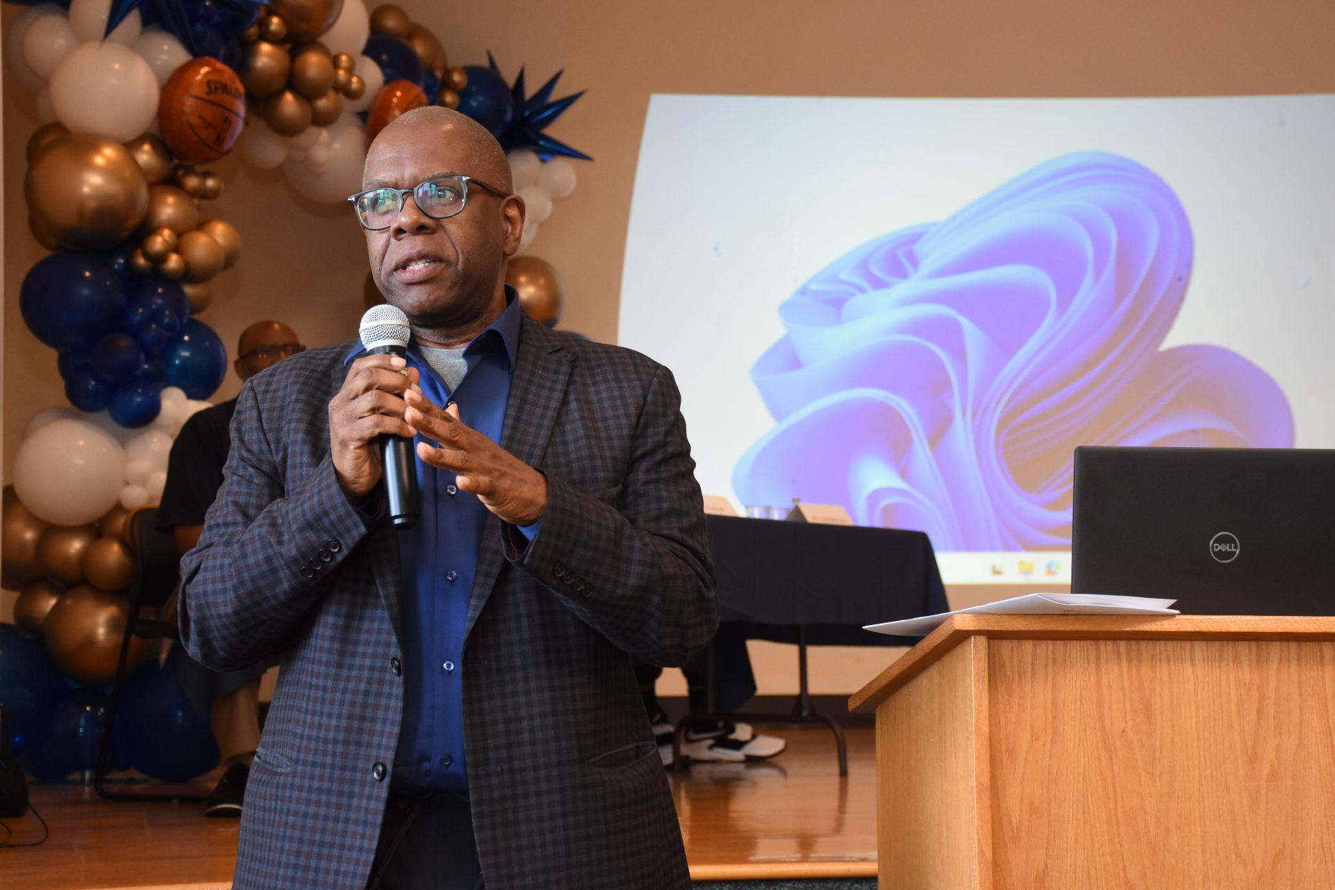 Man speaking into a microphone at an event with balloons and a presentation screen.