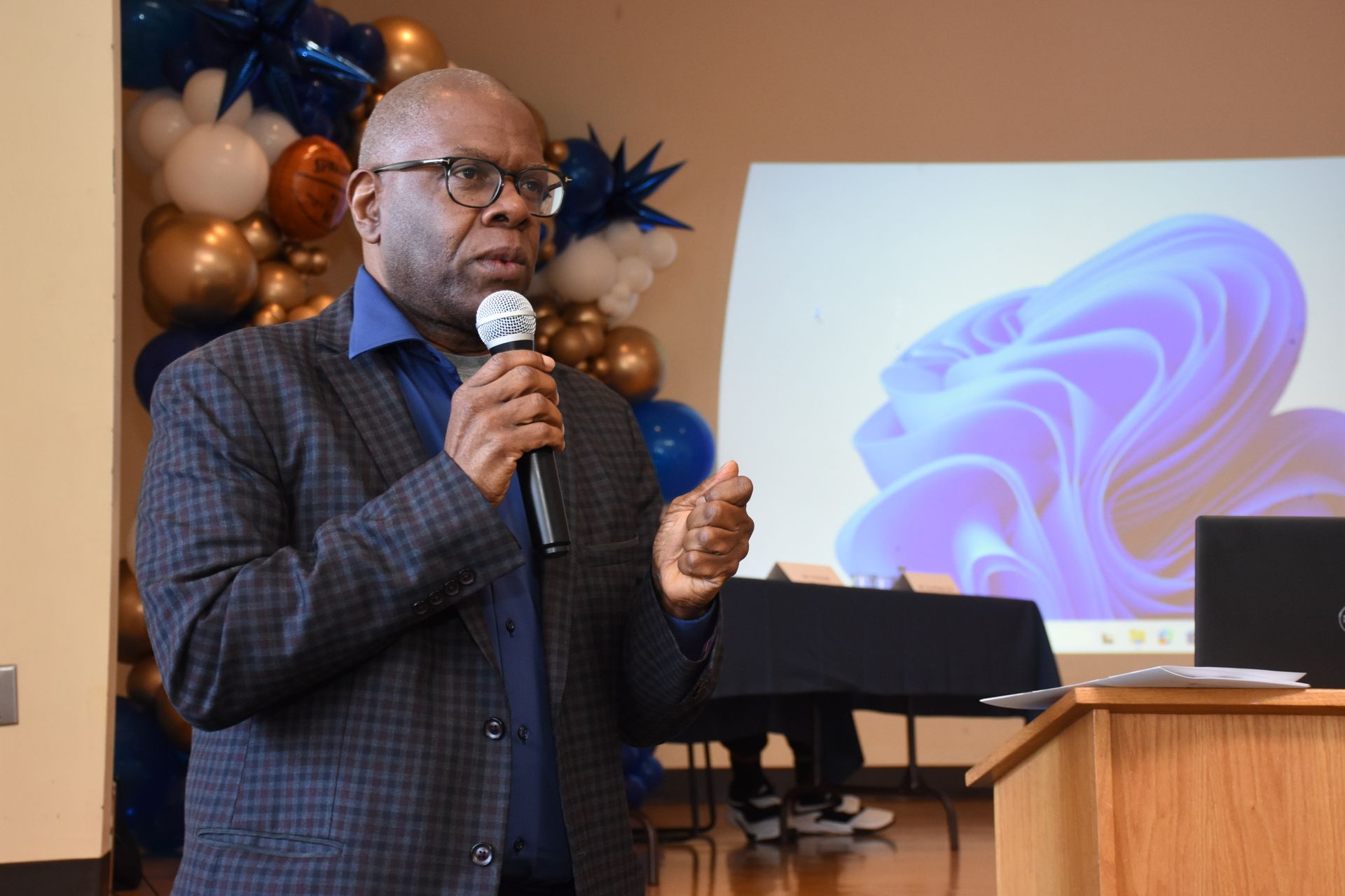Man in a blazer speaks into a microphone at a podium. Balloons and a projected screen are in the background.