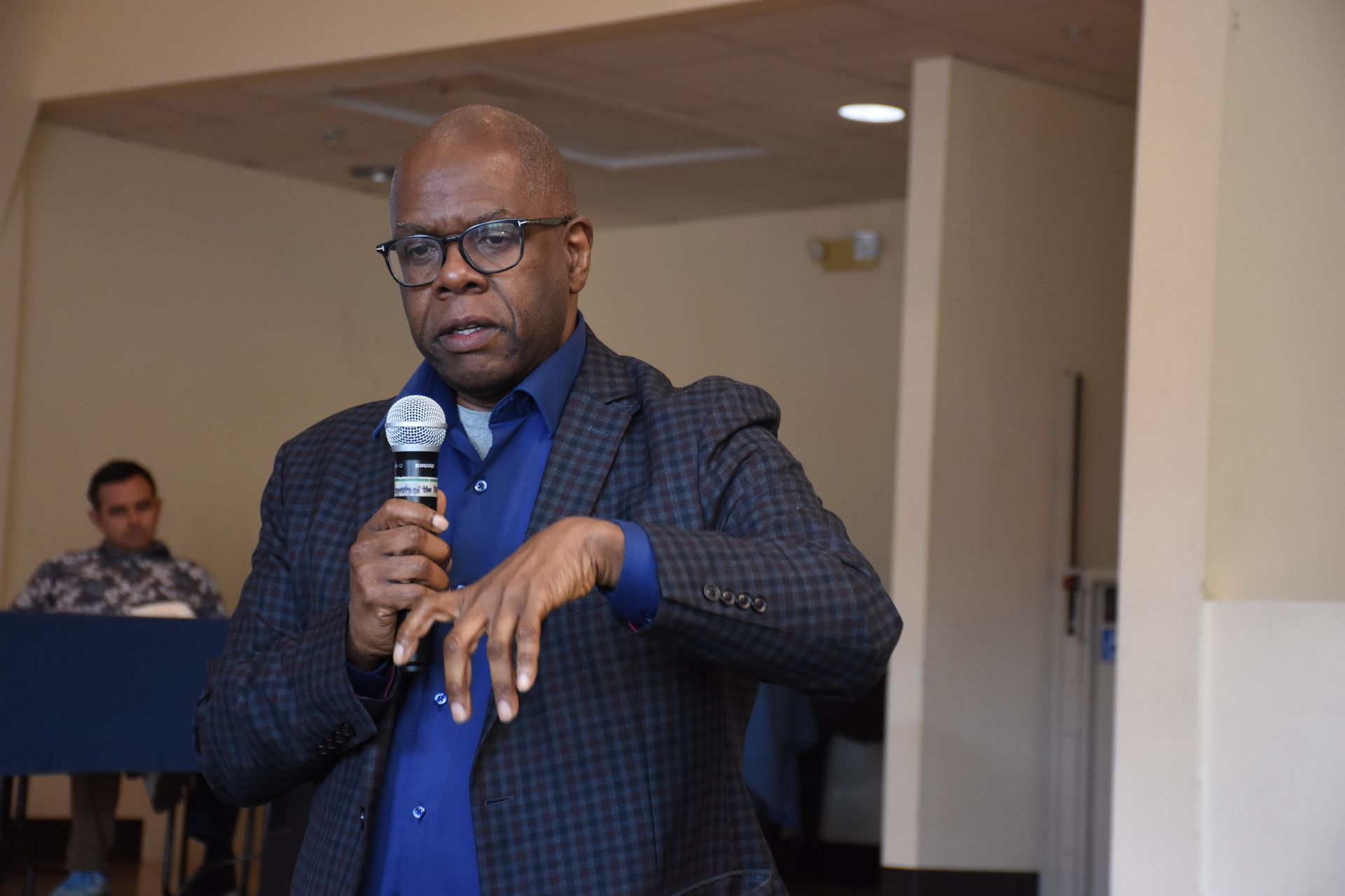 Man in glasses speaks into a microphone, gesturing with his hands. Indoor setting, light walls.