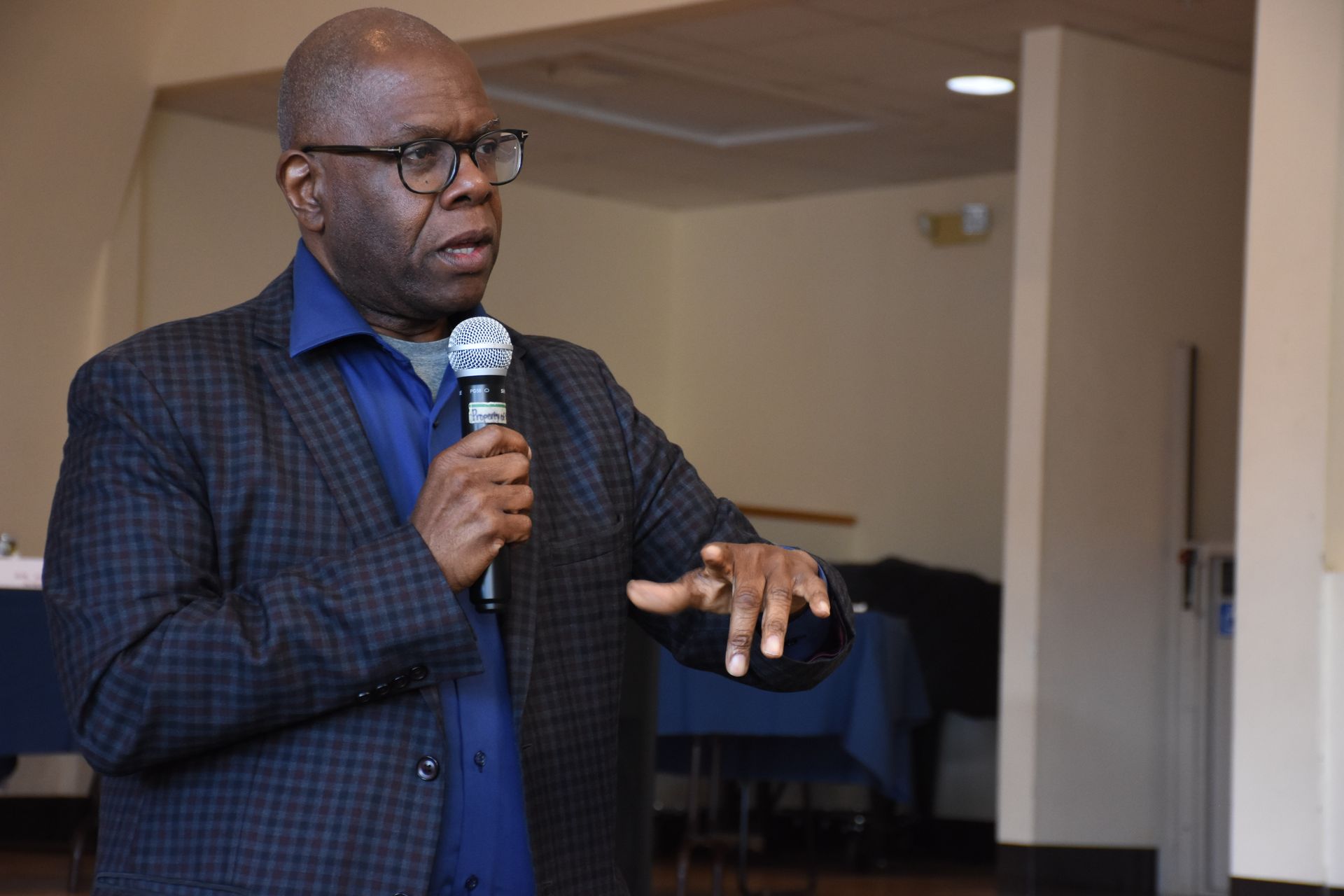 Man in glasses and blazer speaks into microphone, gesturing with his hand.