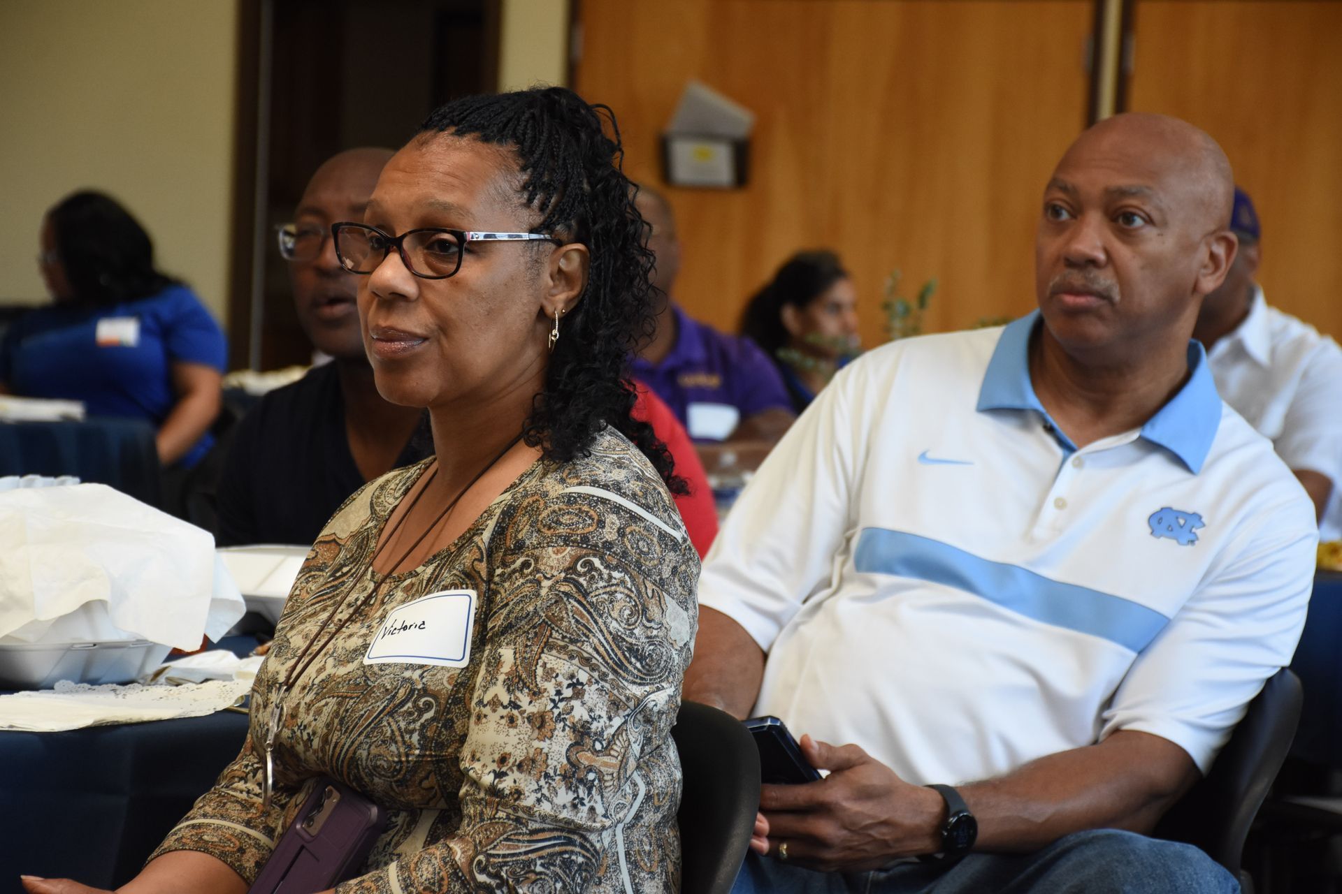 Attendees at a conference. Woman in patterned shirt, man in a white polo shirt, both looking ahead.