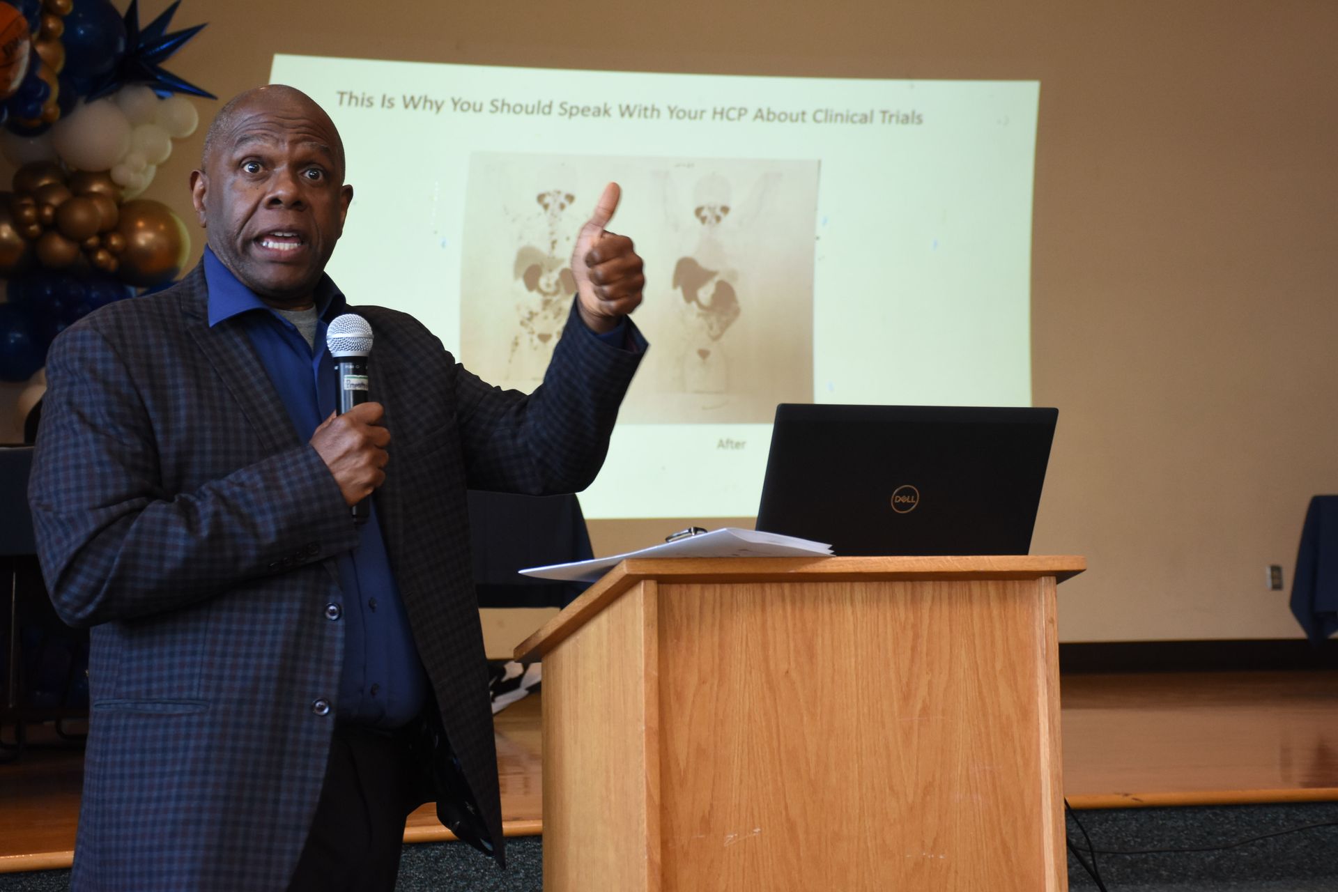Man presenting at a podium, gesturing toward a presentation screen with a thumbs up.