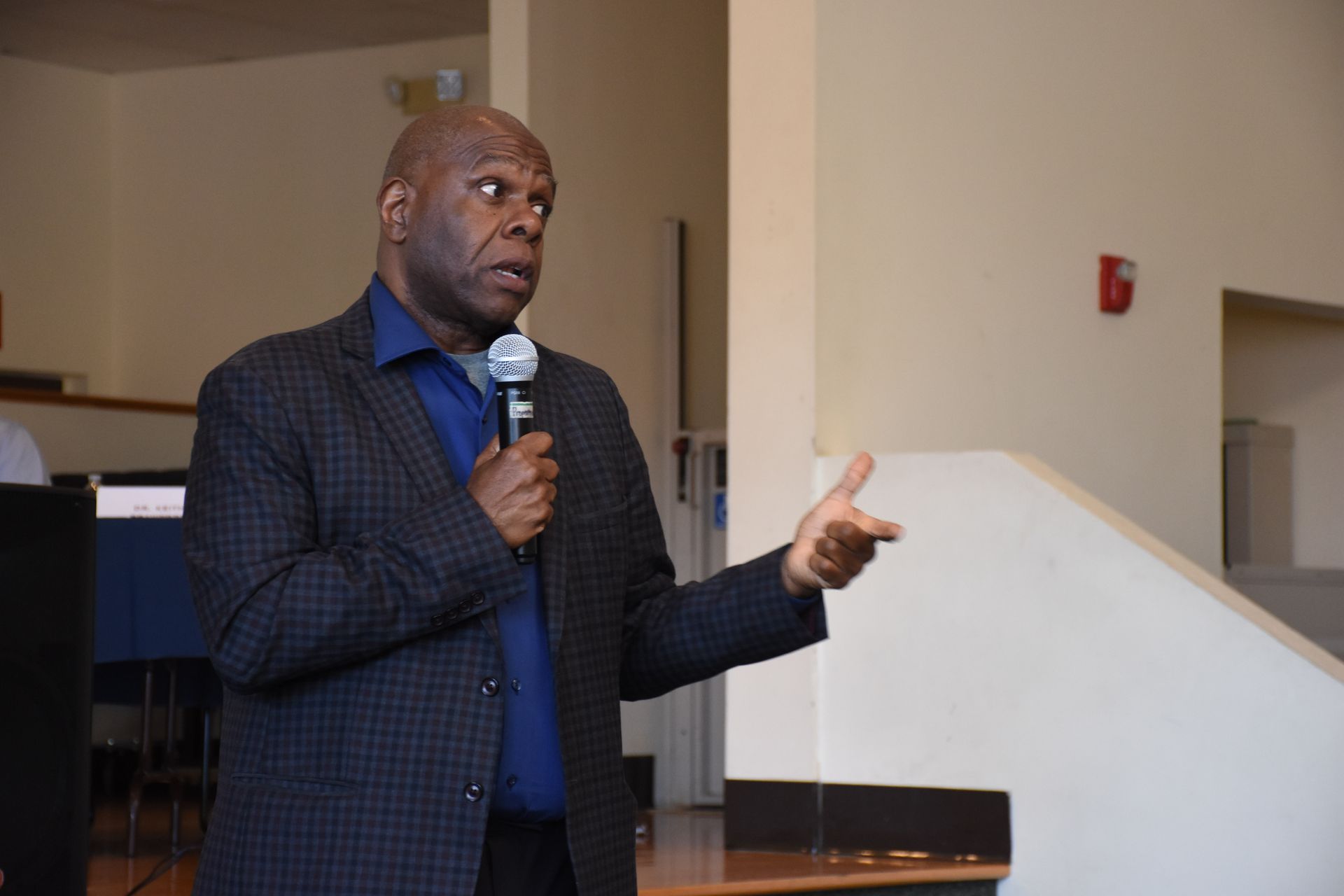 Man in blazer speaks into microphone, gesturing with his right hand in an interior setting.