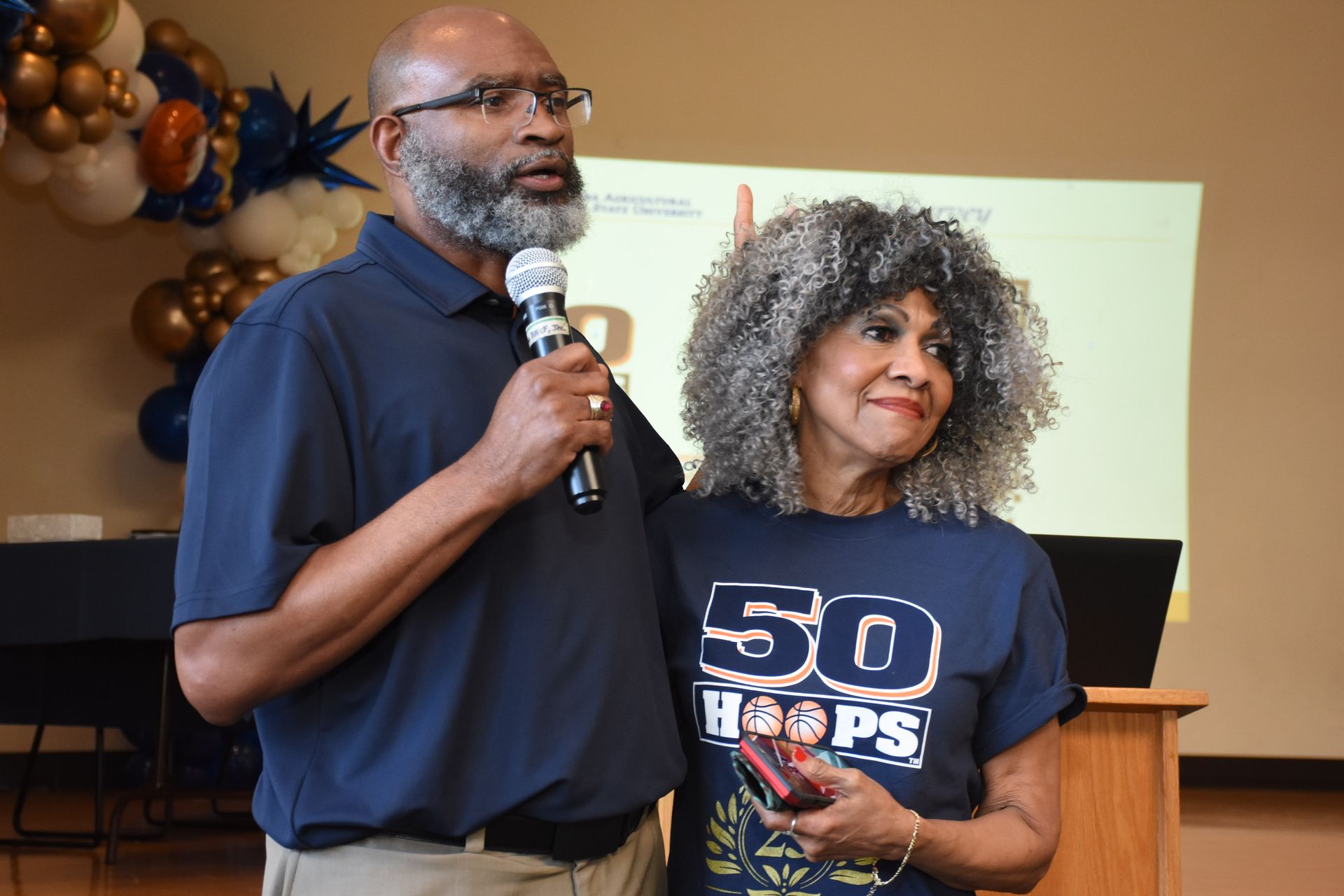 Man speaking into microphone, woman beside him wearing 