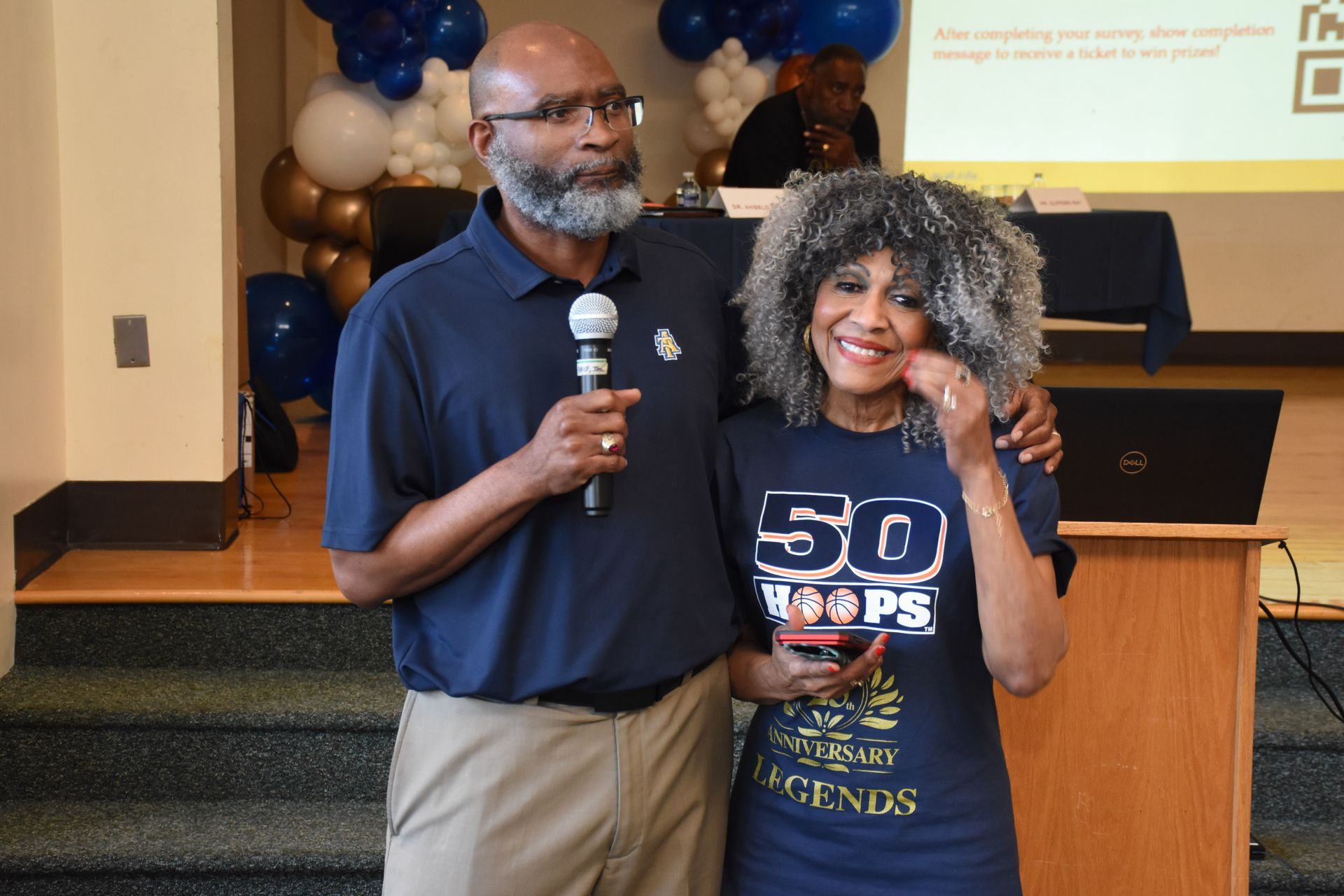 Man with microphone and woman with gray curly hair smiling, arms around each other, stage backdrop.
