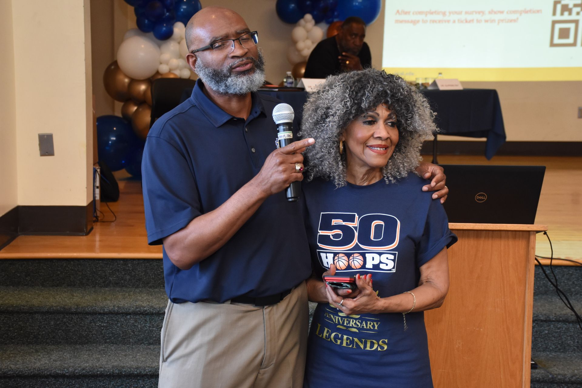 A man with a microphone and a woman with an arm around her, both smiling, at an event.