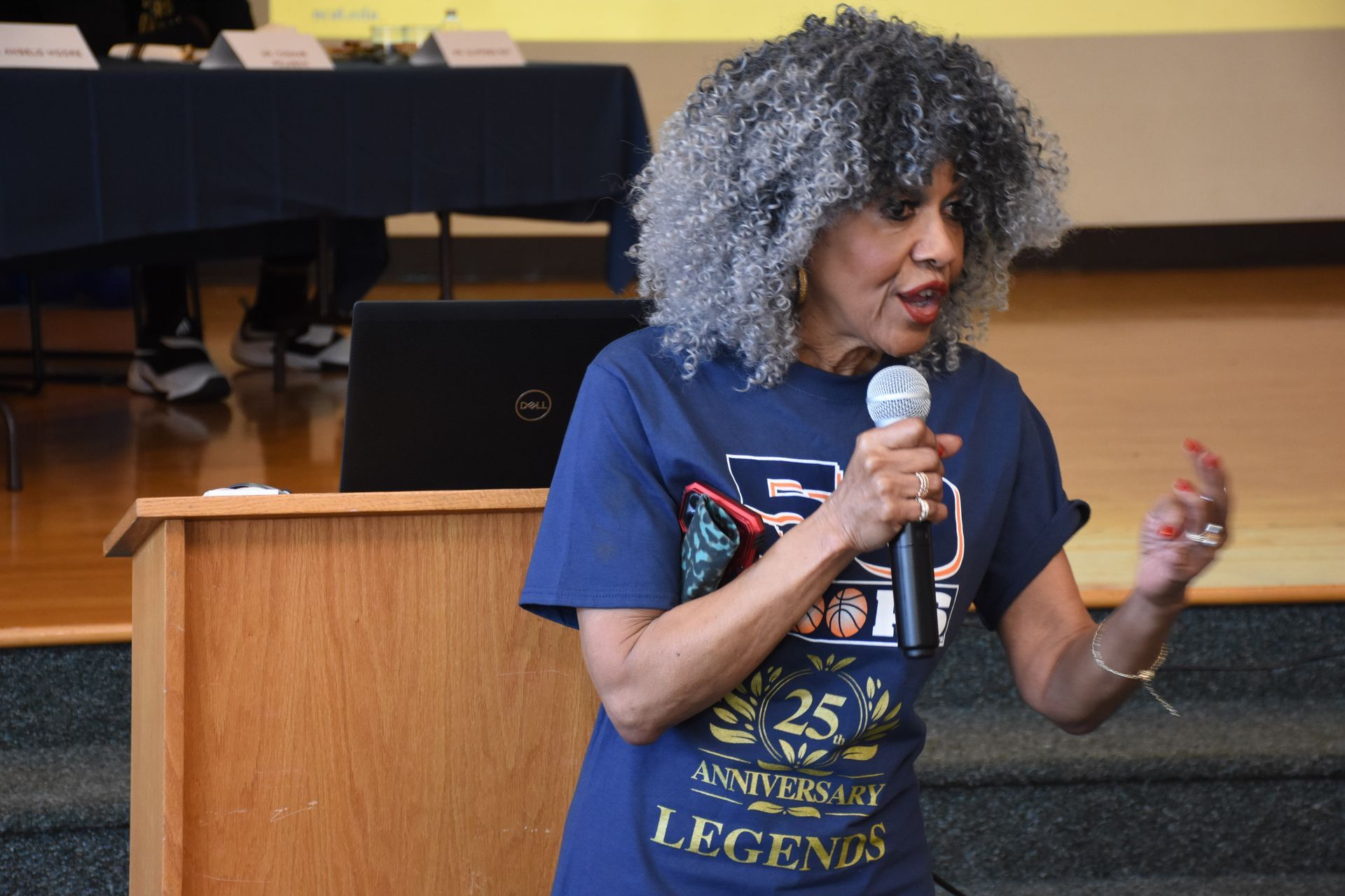 Woman speaking into a microphone at a podium. She wears a navy blue t-shirt and has gray hair. Indoors.