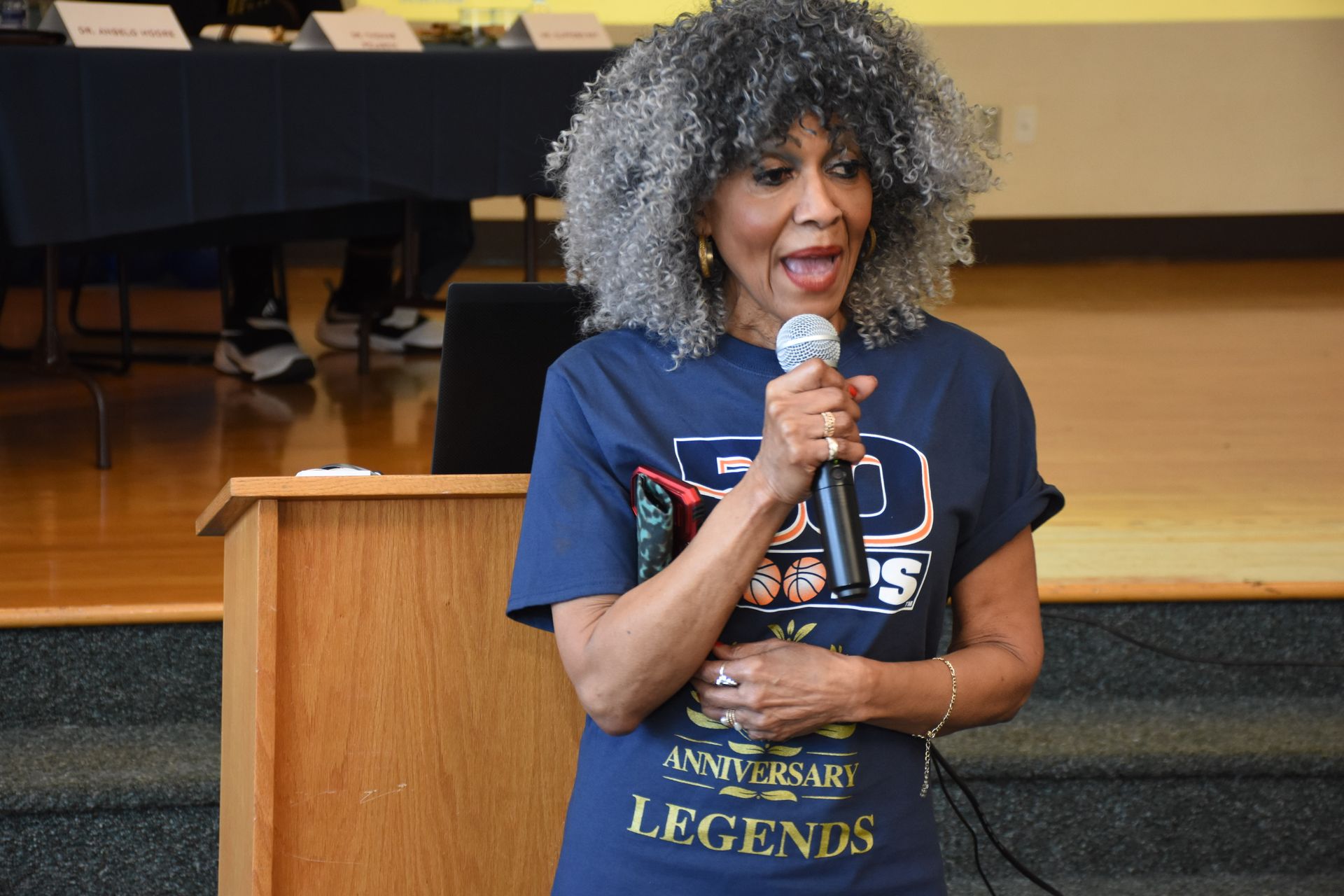 Woman with gray curly hair speaks into a microphone from a podium; 