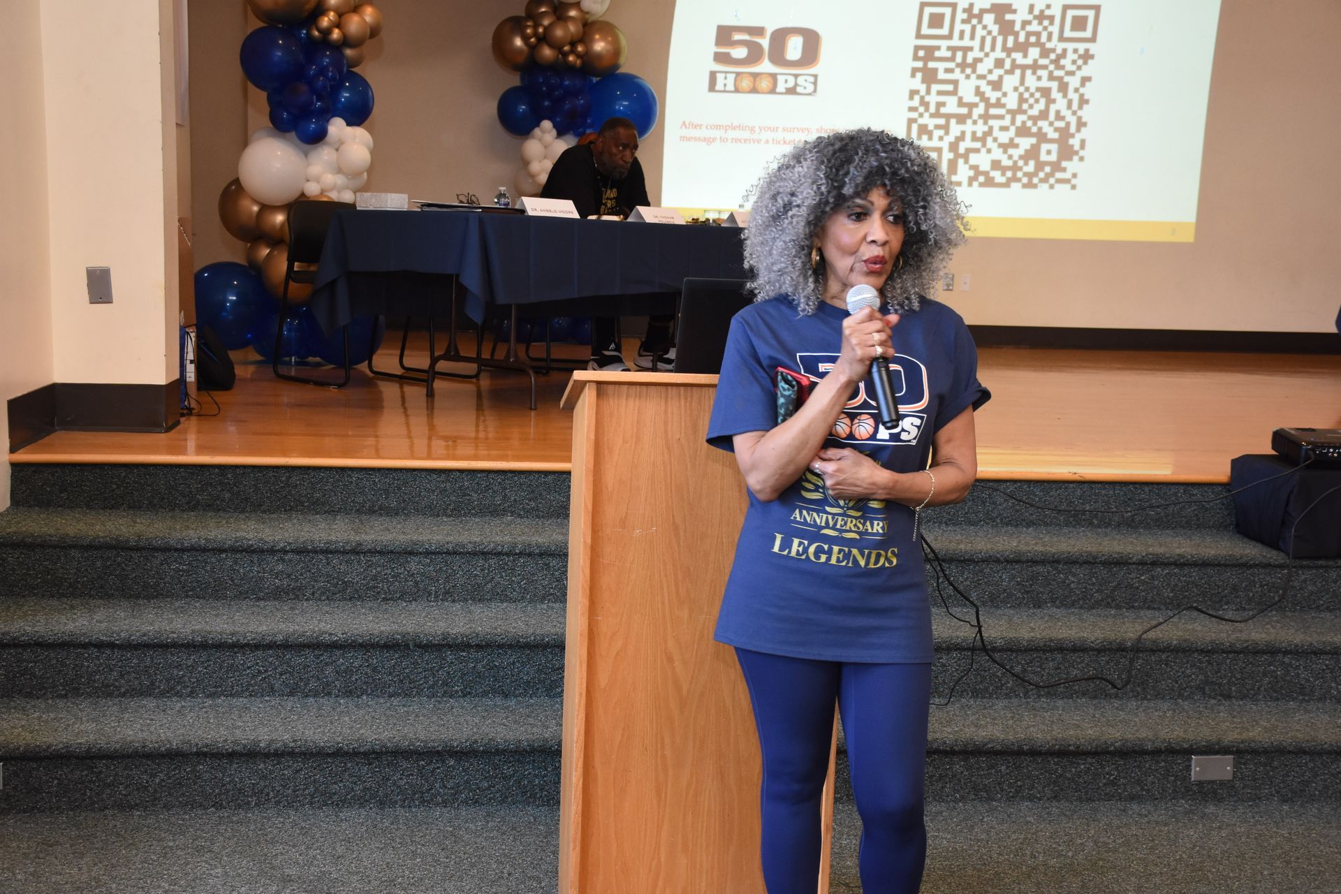 Woman speaking at a podium, wearing blue shirt and leggings. Balloons and a screen behind.