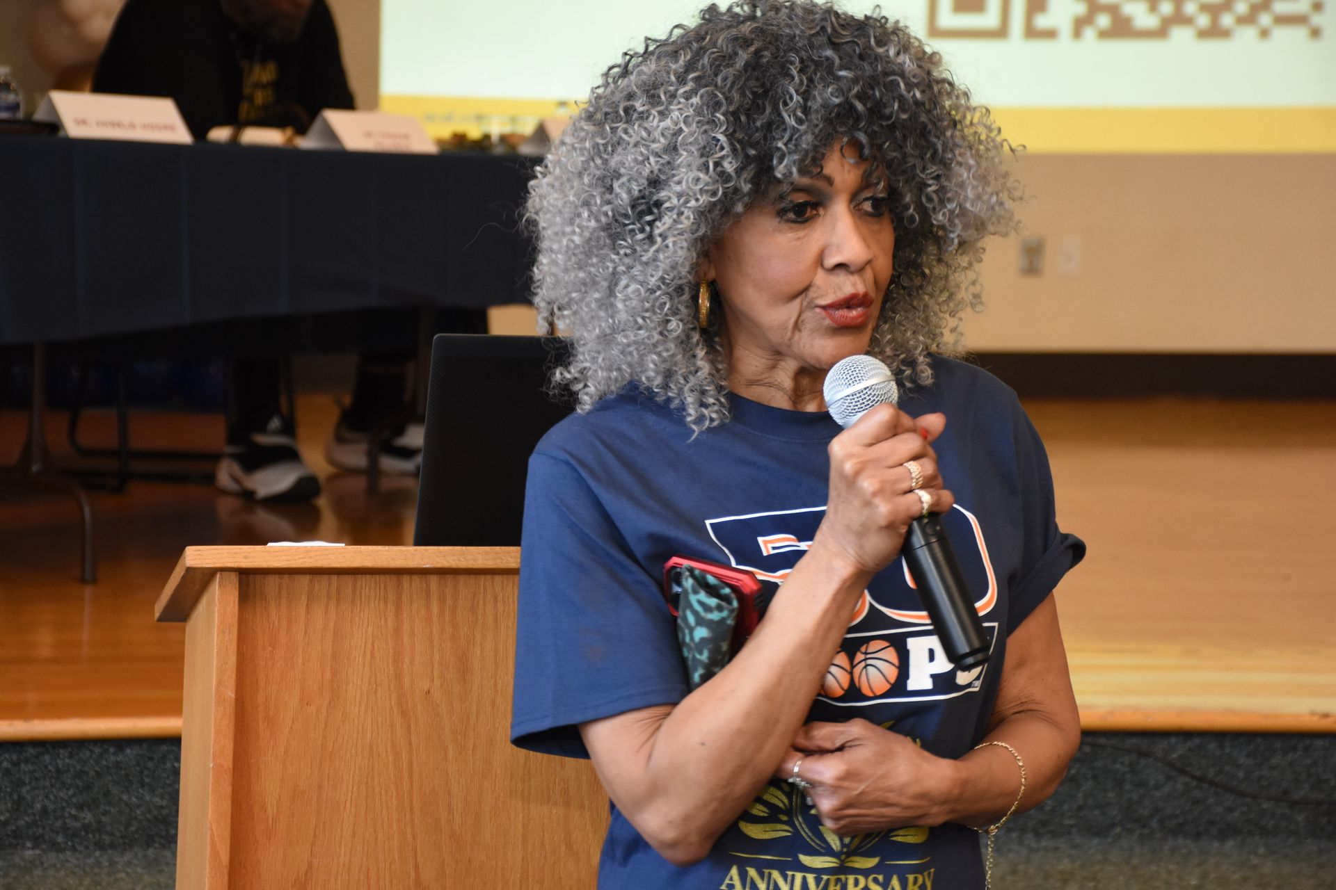Woman with gray afro speaks into a microphone at a podium. She wears a blue shirt and holds notes.
