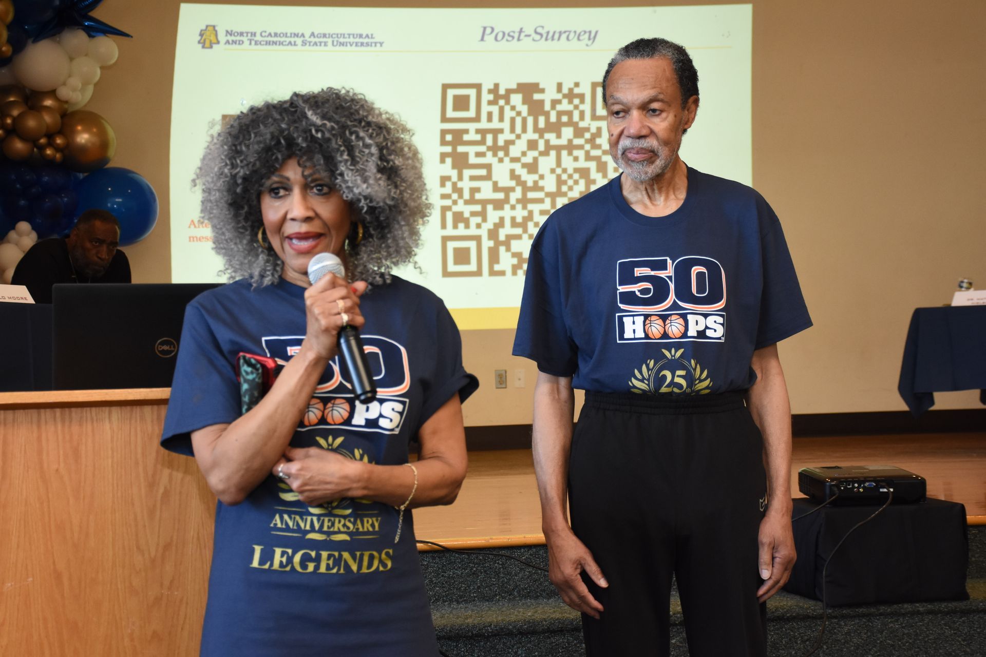 Two people in matching t-shirts with microphones, standing in front of a banner.