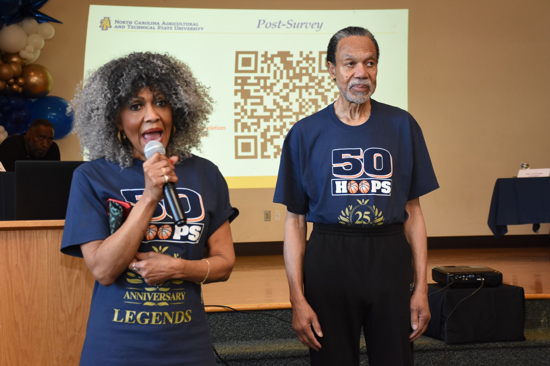 Two people in matching t-shirts stand at a podium. A woman speaks into a microphone, while a man stands beside her.