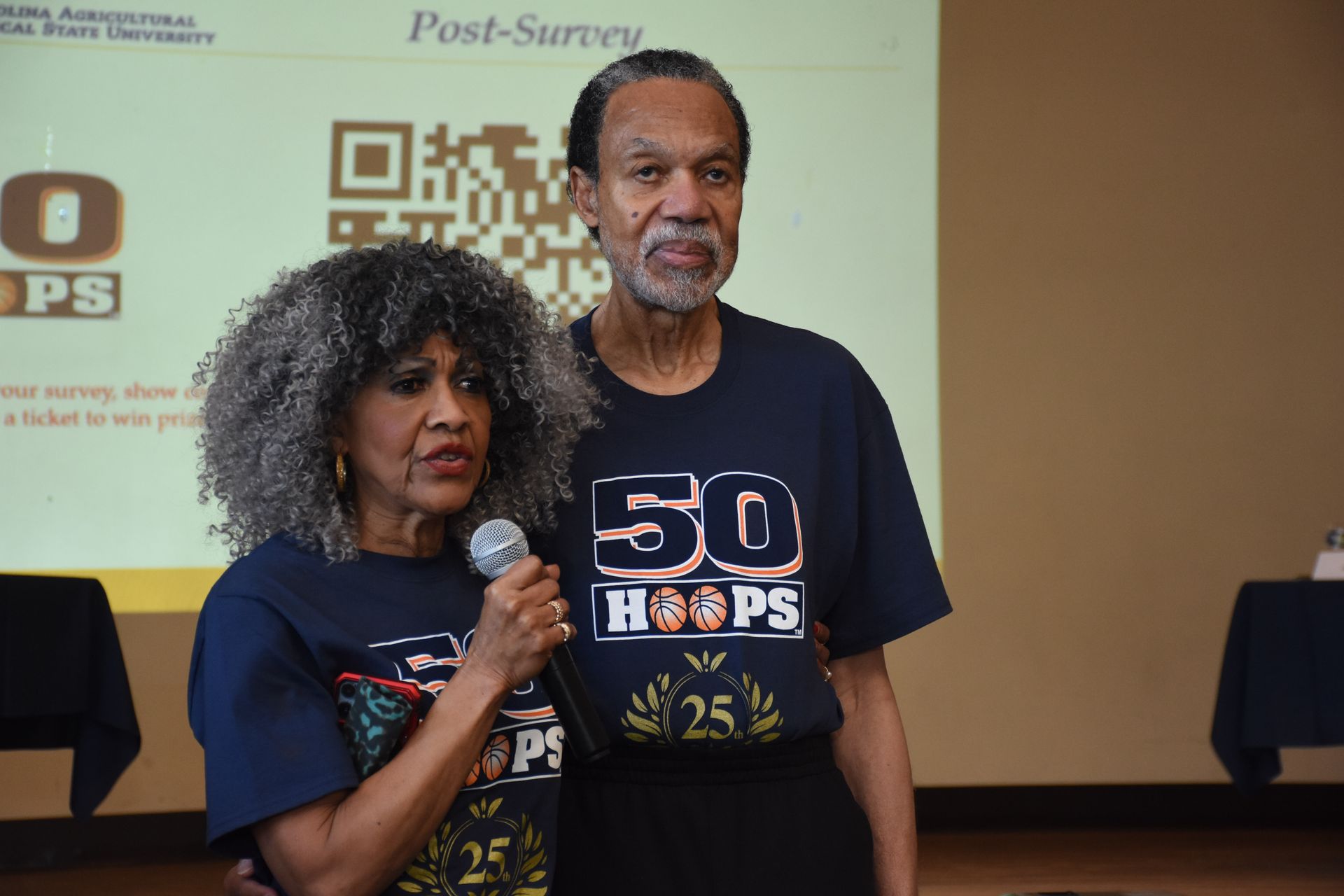 Two people in matching shirts stand in front of a projection screen. They are holding a microphone.