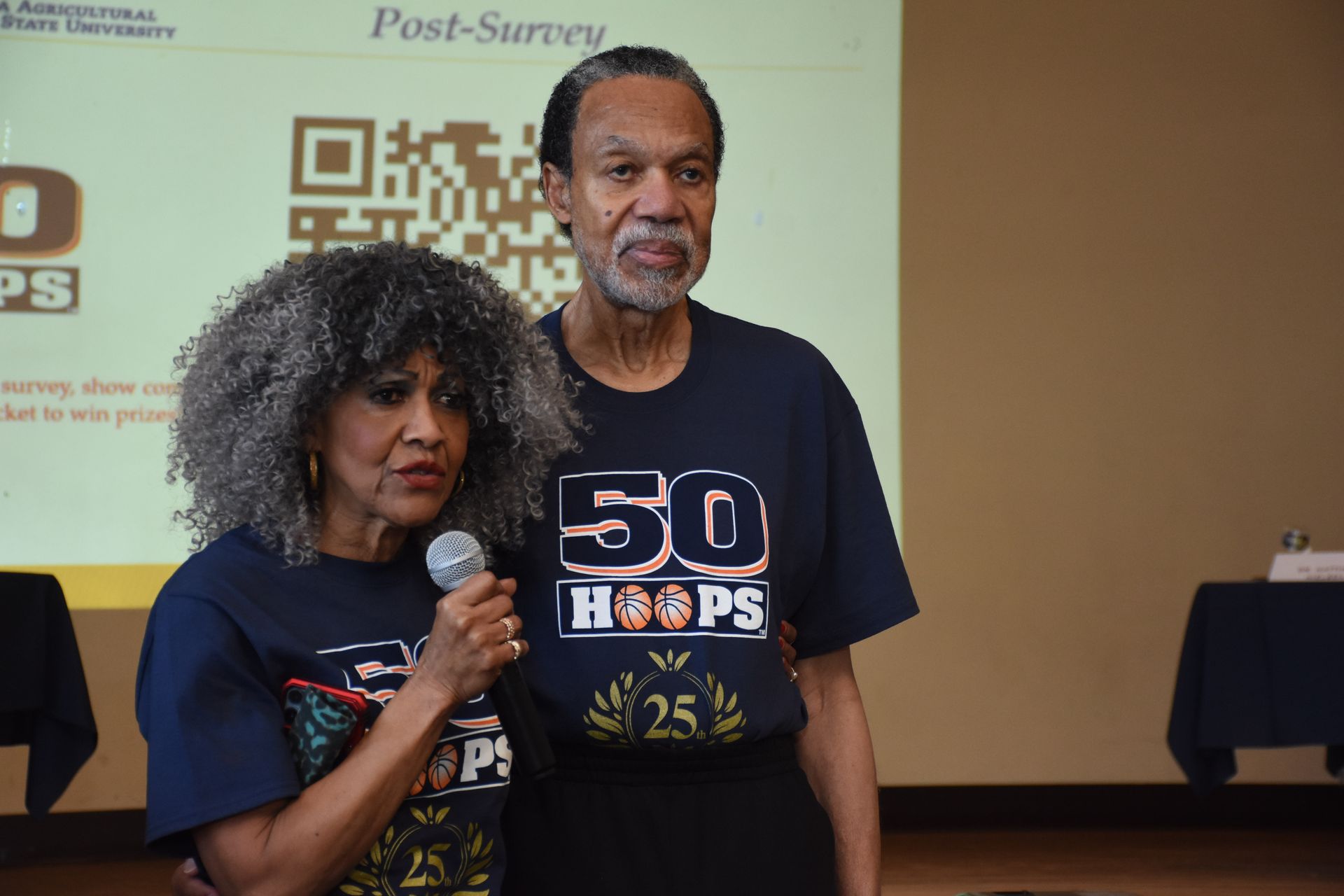 Two people on stage, holding microphone, wearing matching 