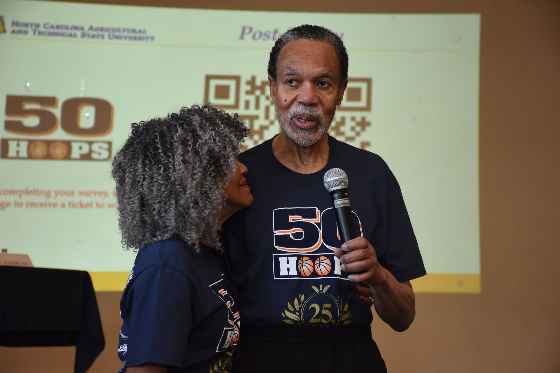 Man speaking into a microphone, standing next to a woman; both wearing 