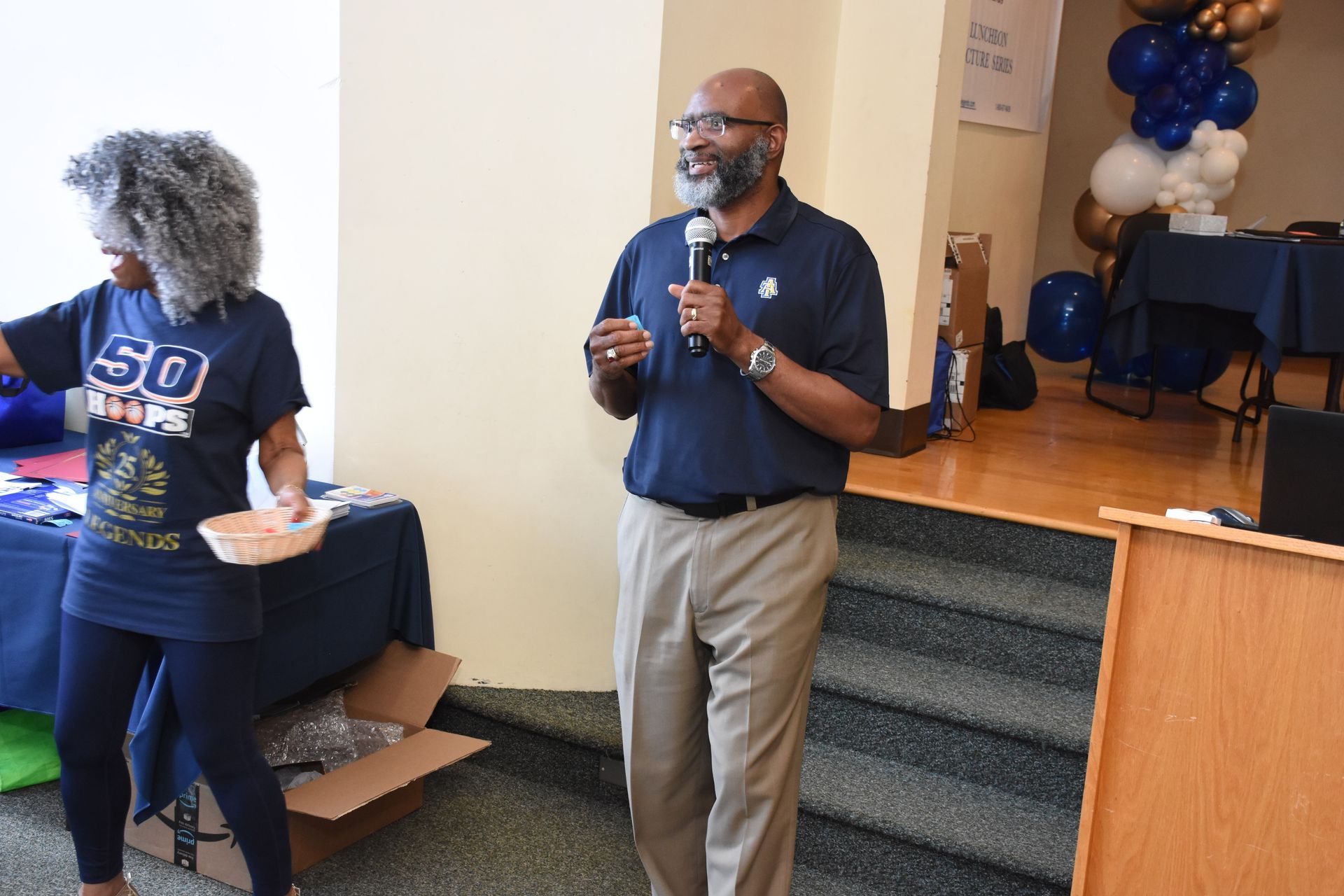 Man speaking into microphone on stage; woman nearby, setting with balloons and table.