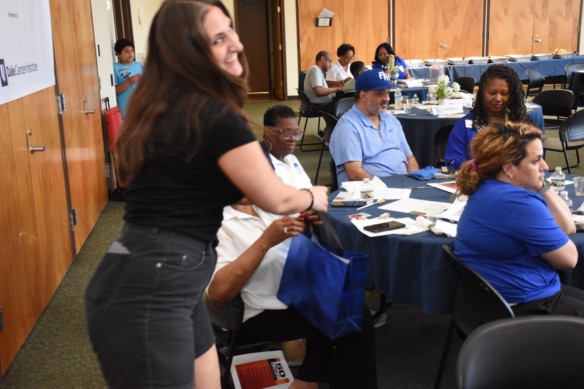 Woman reaching toward a seated person at a table in a conference room; other attendees are present.