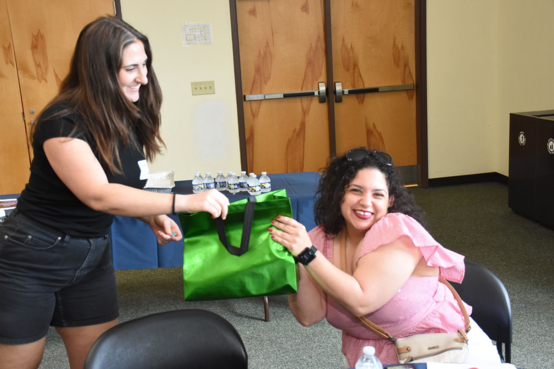 Woman hands a green bag to a seated woman who smiles and claps at a table in a room.
