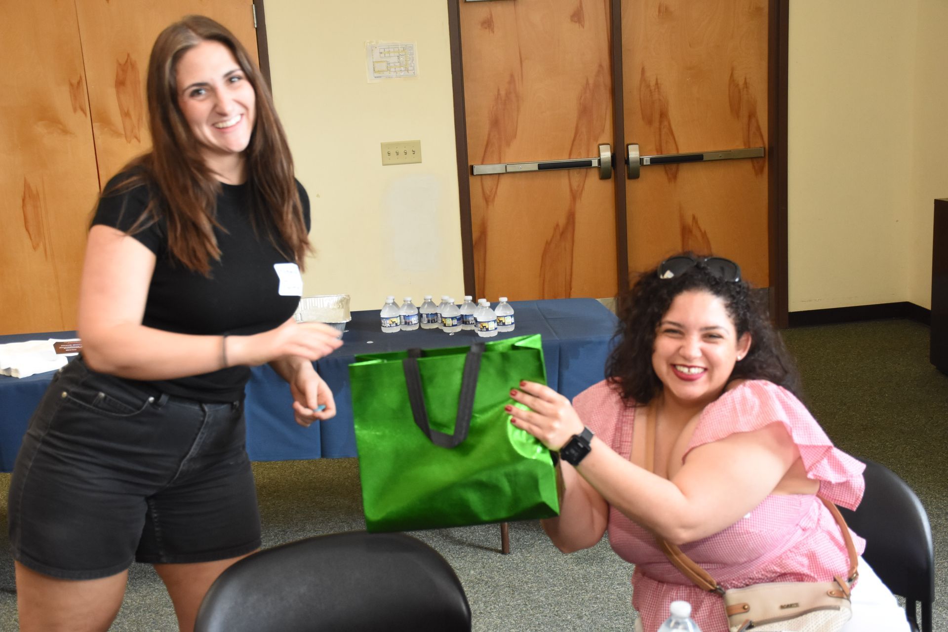 Two women smiling, one holding a green bag, the other holding small objects, in a room with wood paneling.