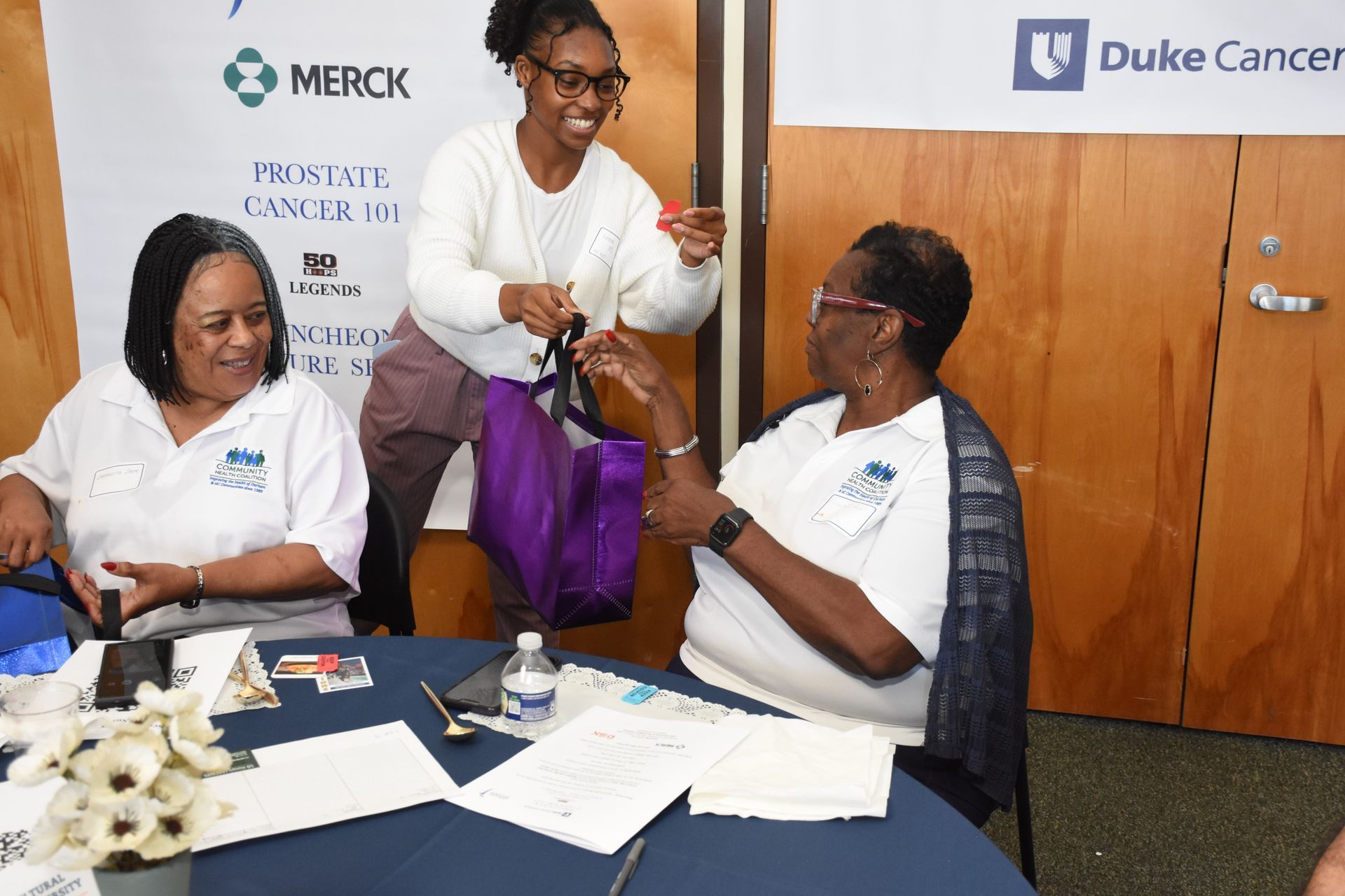 Woman giving a purple bag to another woman at a table with papers and flowers; Duke Cancer event.