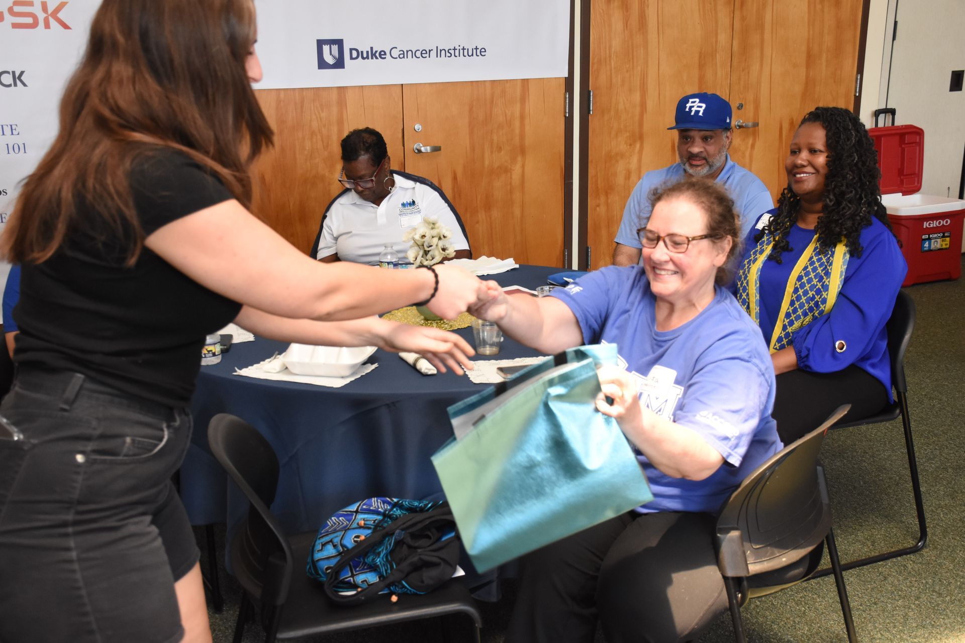 Woman hands a gift bag to a woman seated at a table, smiling. Two others seated nearby. Indoor event.