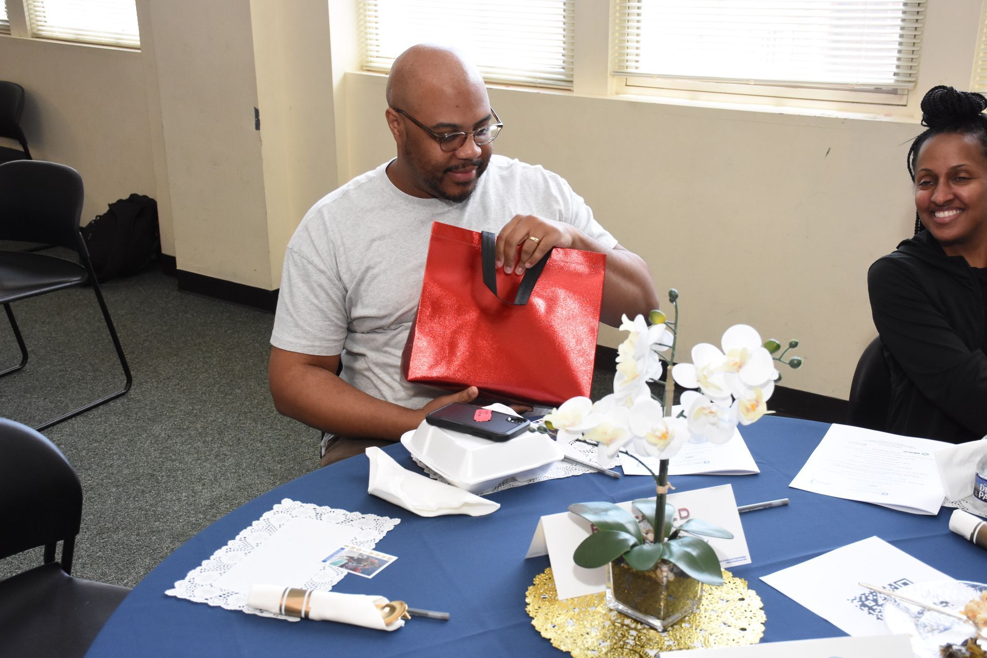 Man opening a red gift bag at a table set for an event; woman smiles nearby.