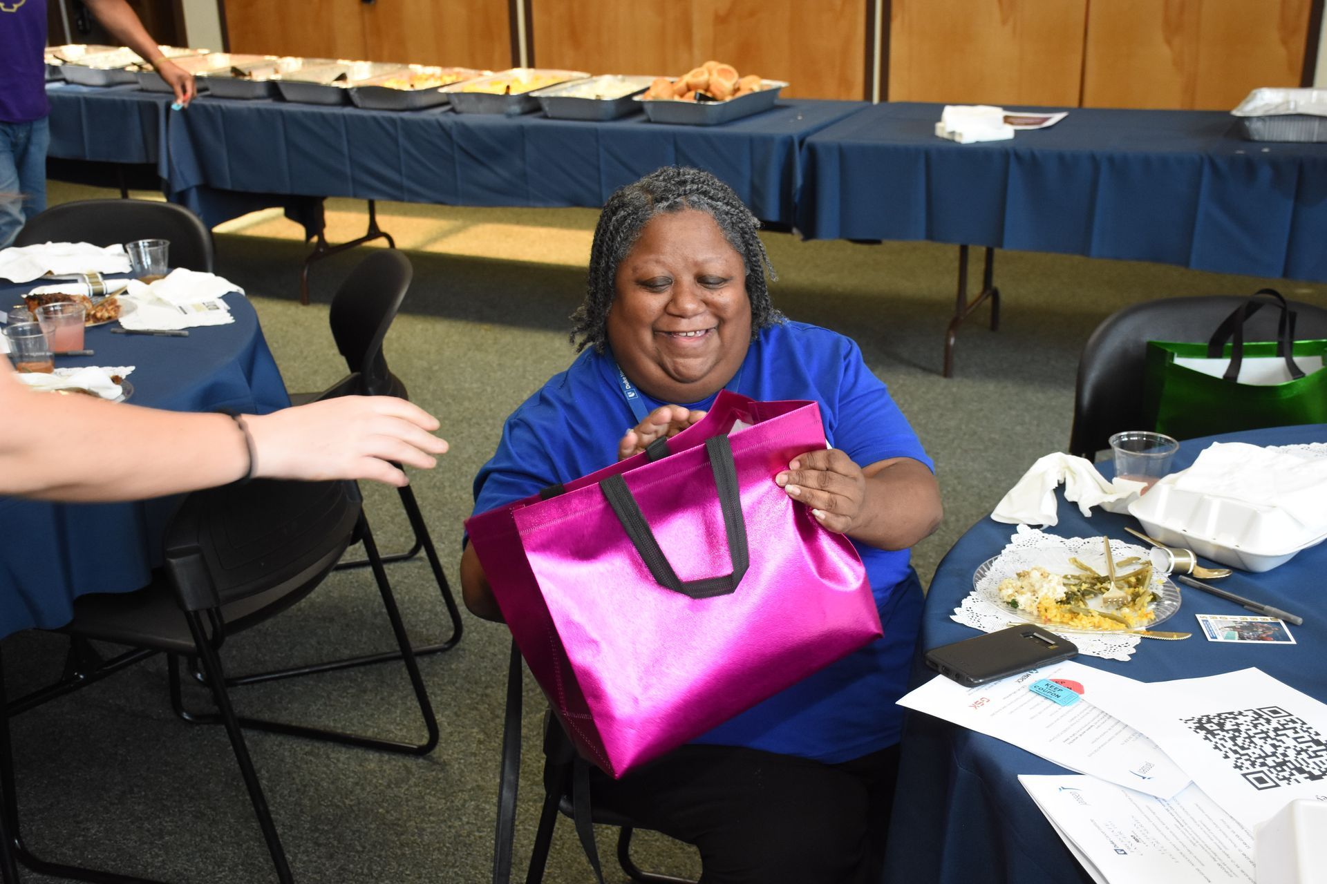 Woman in blue shirt smiles, holding a pink gift bag at a table. Food on tables in background.