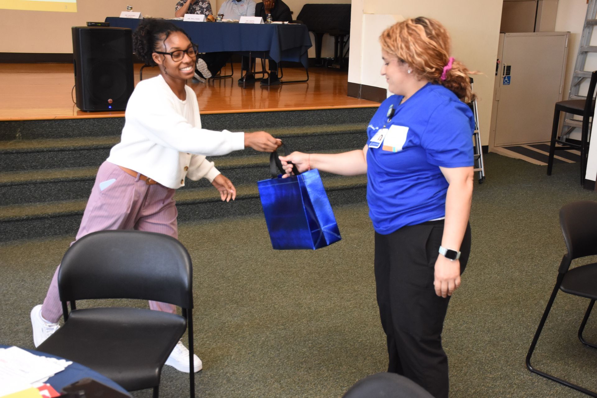 Woman in blue shirt gives gift bag to another smiling woman. Interior setting, audience.