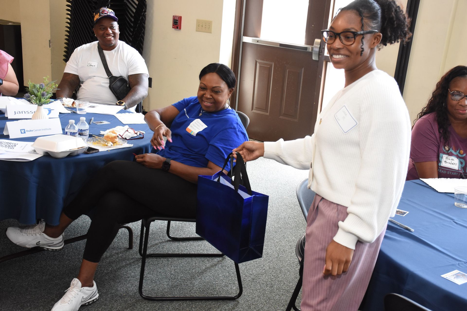 Person handing a blue tote bag to another person at a table; others seated nearby.