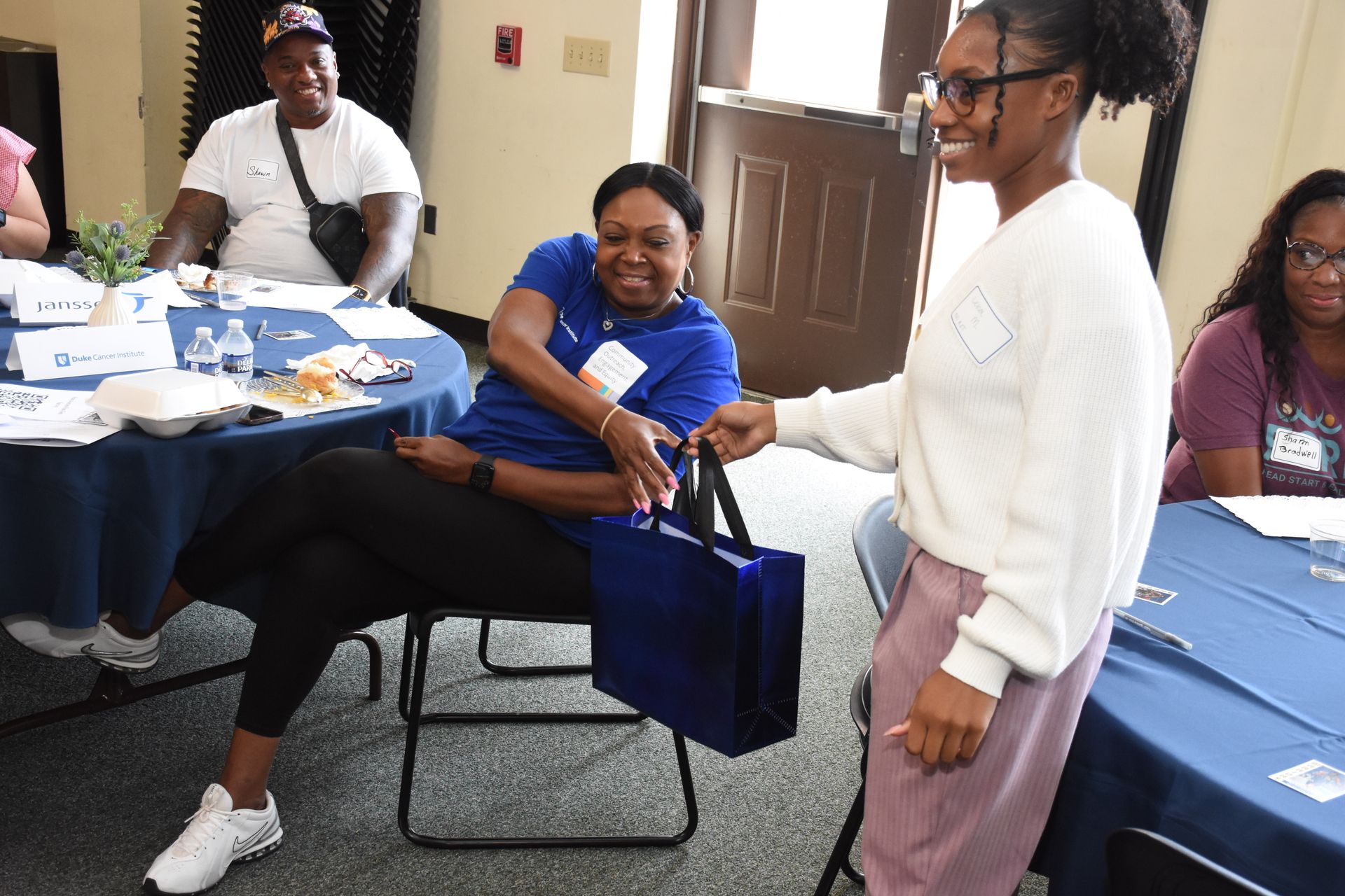 Woman in blue shirt receives a blue gift bag from a person in a white sweater at a table with others.