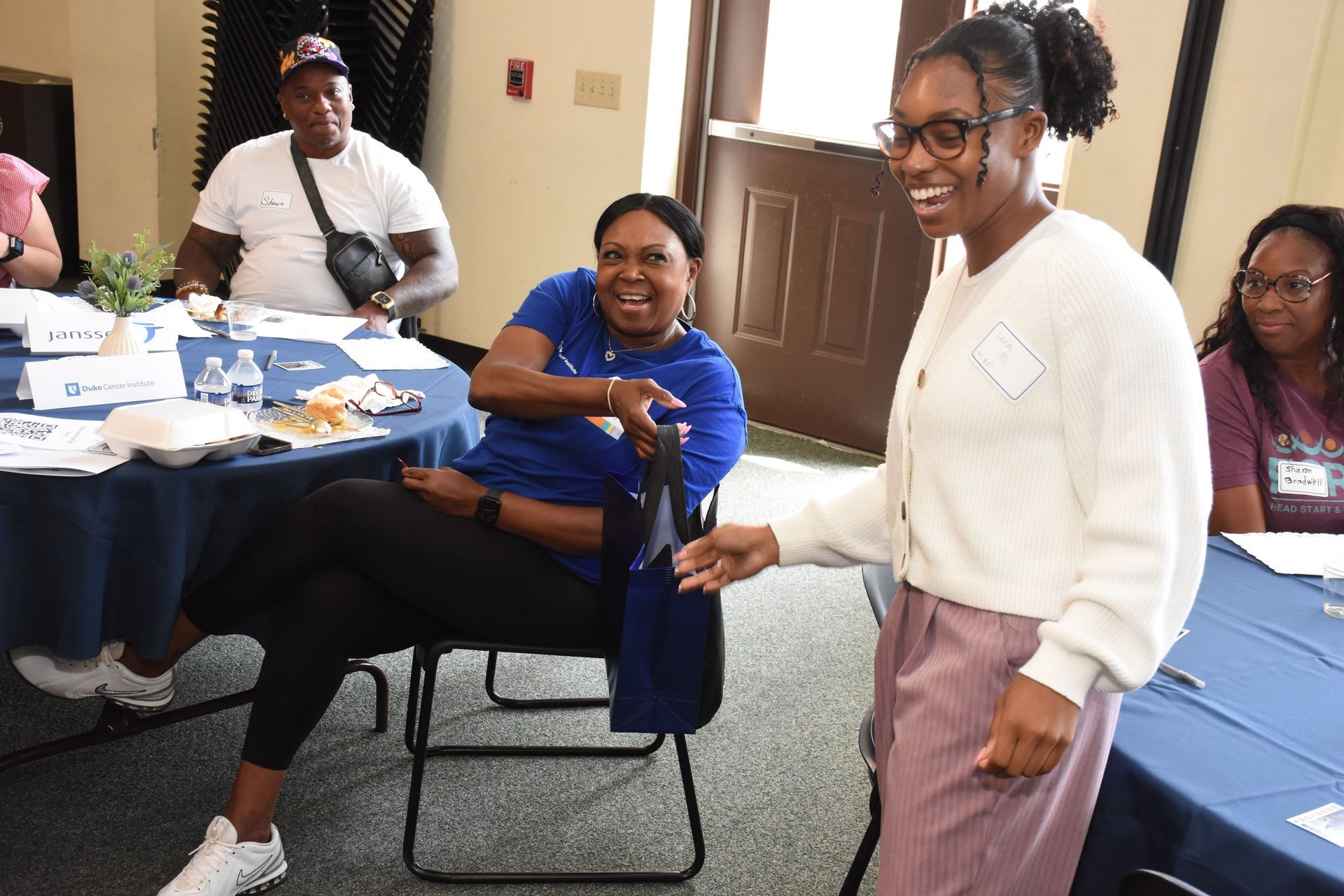 People laughing at a table in a room. One stands, holding something. Others are seated.