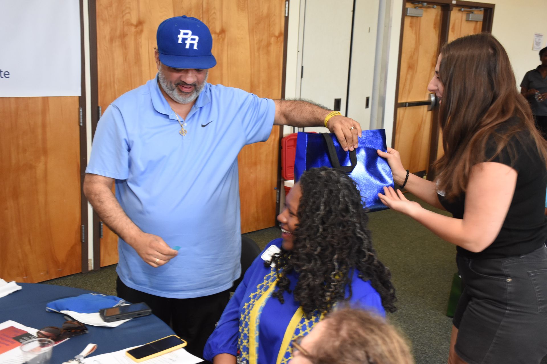 Man in blue cap/shirt gives gift bag to a woman in a blue shirt; another woman looks on. Interior.