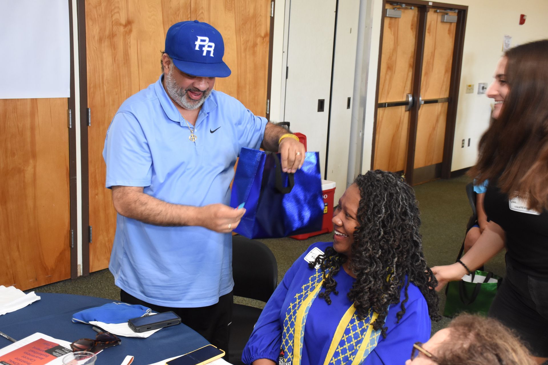 Man in blue shirt and cap shows something from bag to smiling woman. Another woman smiles nearby.