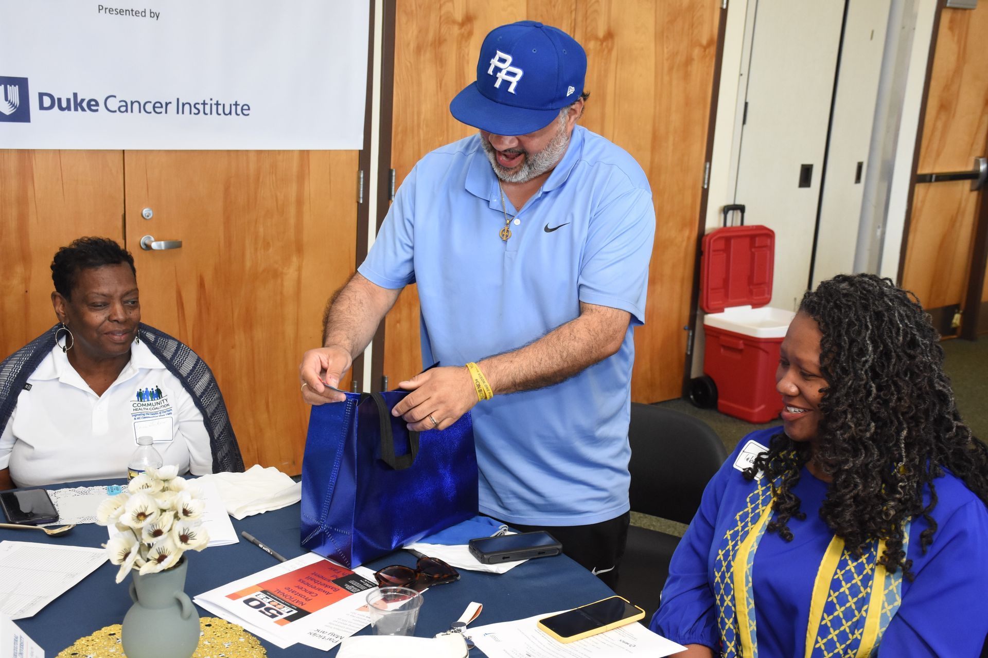 Man handing out a blue bag at a table with two other people; Duke Cancer Institute banner.