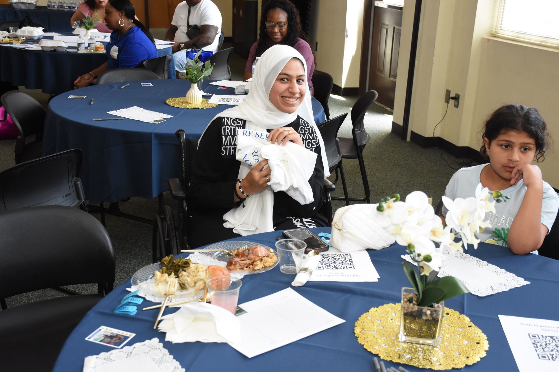 People seated at tables during an event, some smiling, with food and floral centerpieces.