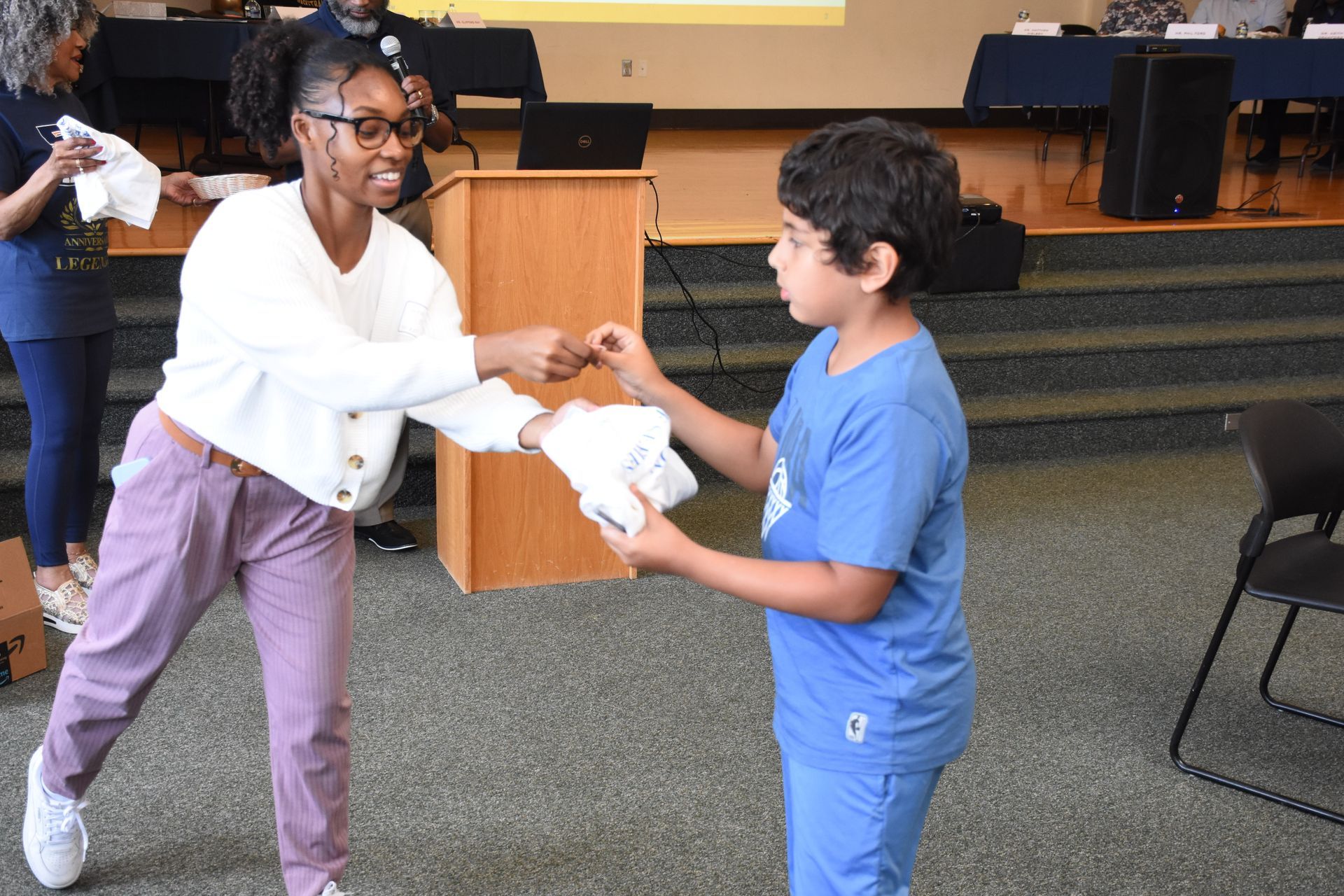 Woman hands a white item to a child; they're in a room with a podium, stage, and chairs.