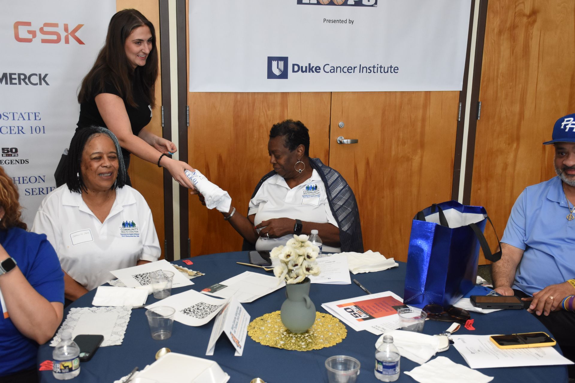 Woman hands papers to people seated at a table, likely an event or meeting. Blue and white colors are present.