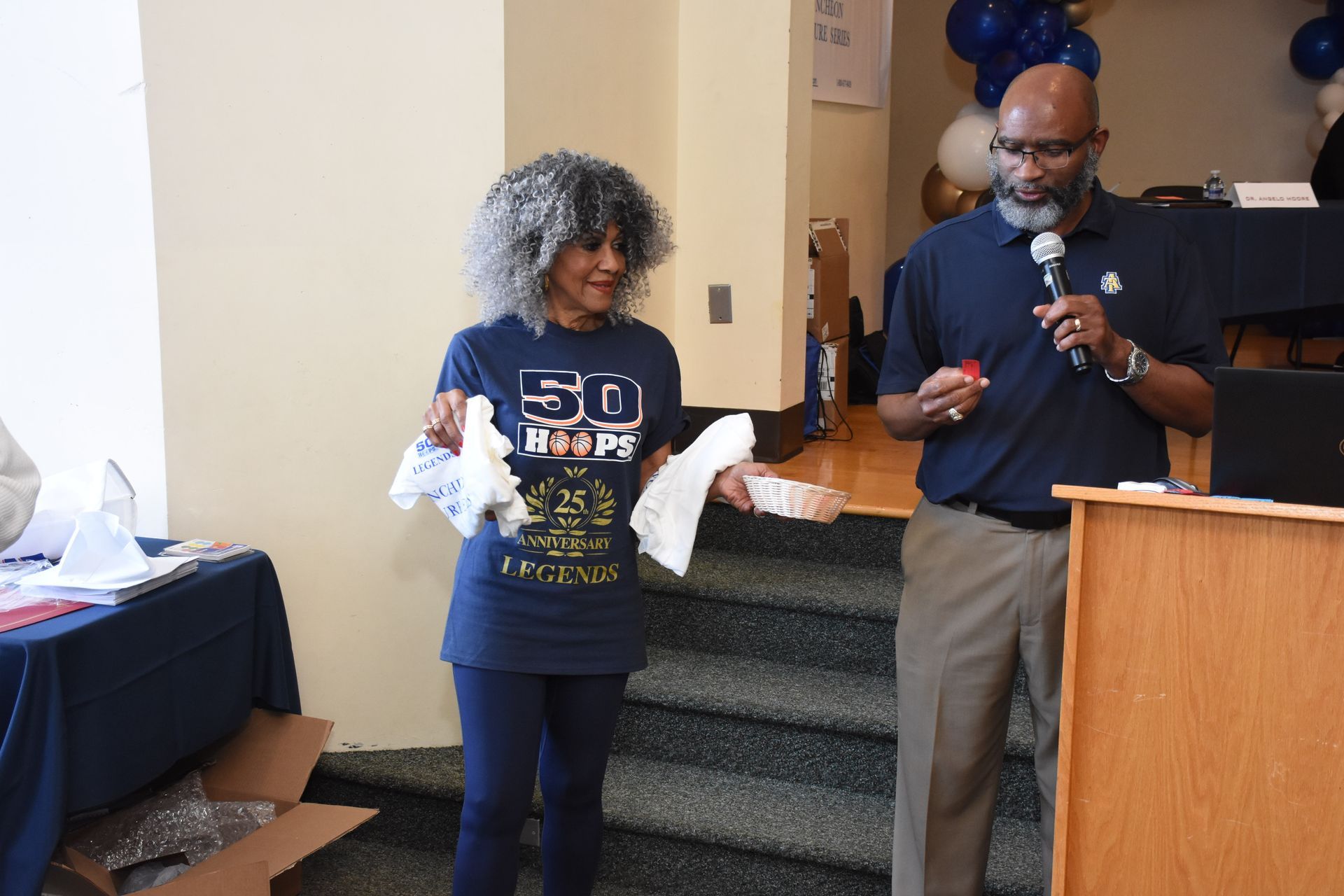 Woman with curly hair and man with a microphone at a podium; holding food items, blue and gold balloons in the background.