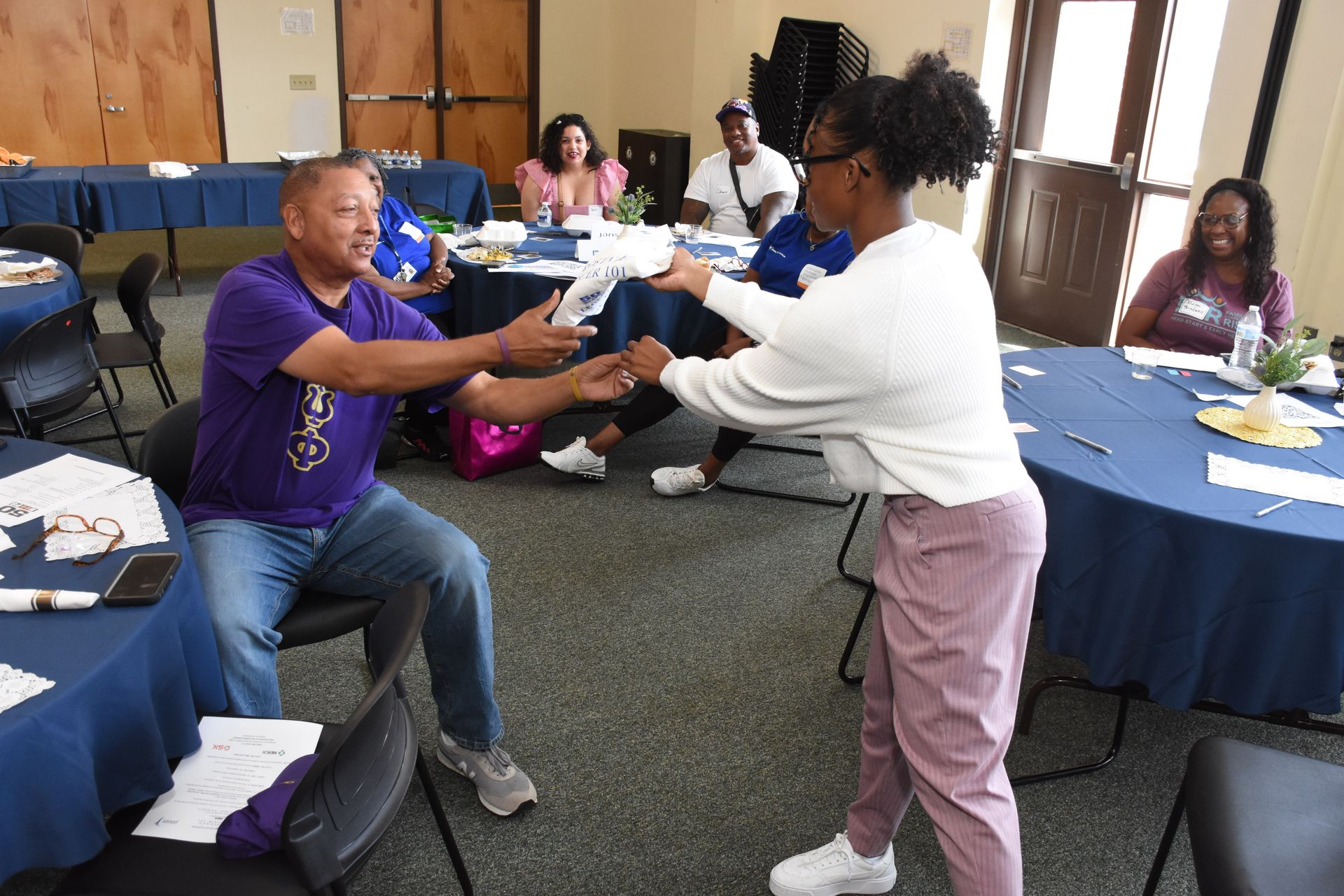 Man and woman passing paperwork during a meeting; others seated at tables.