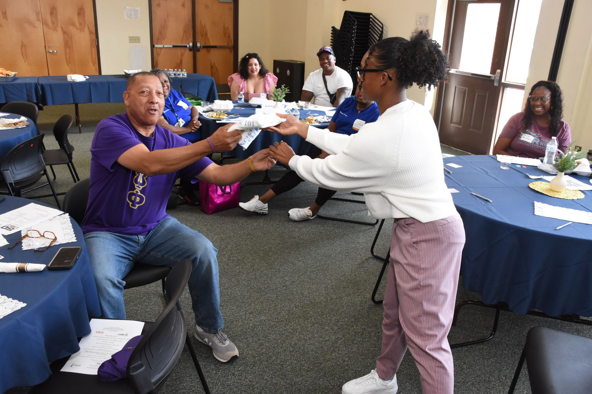 A person hands a paper to another person at a table, people are seated at round tables in a room.
