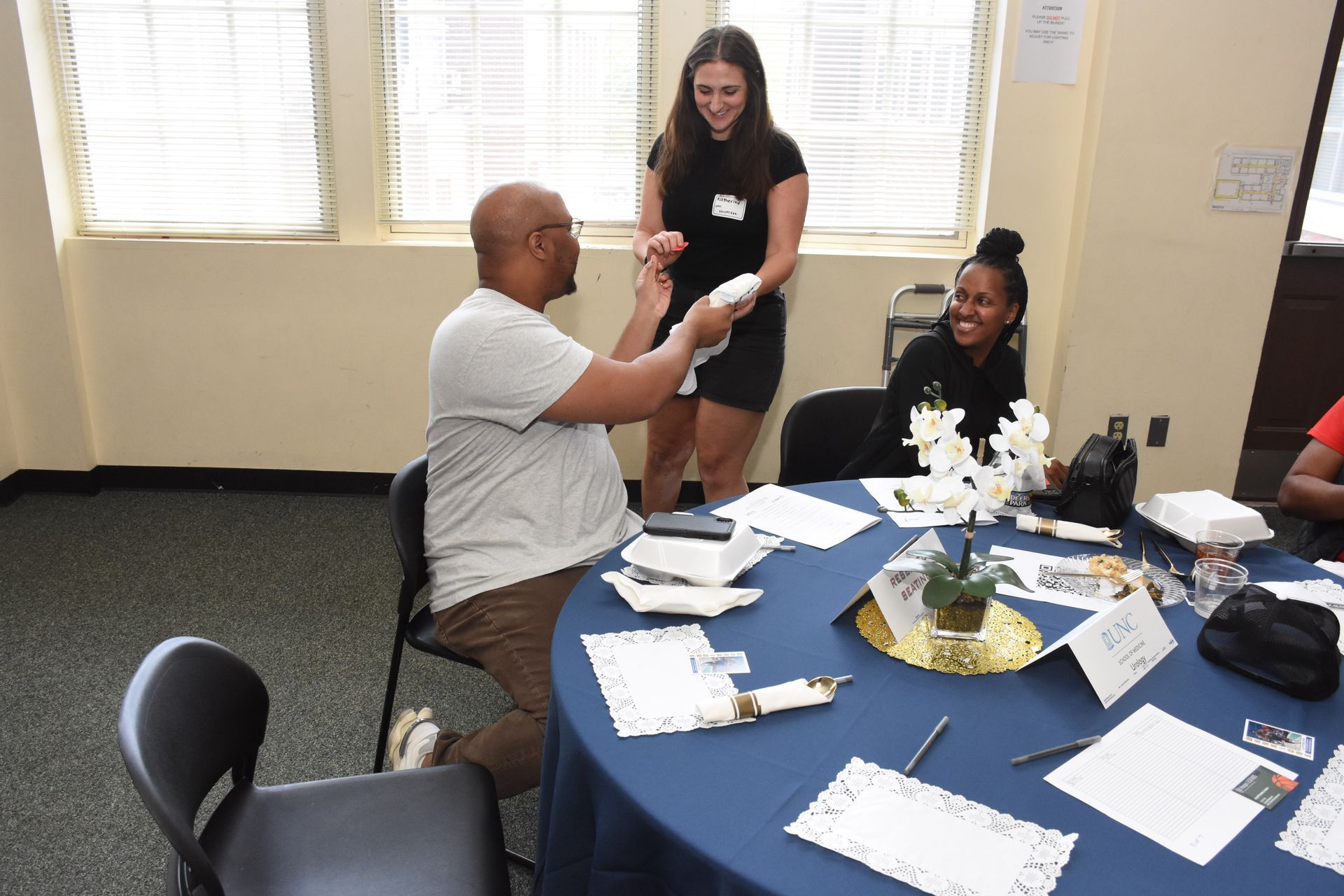 People at a table. A man seated, holding items. A woman smiles, handing him something. Another woman at the table smiles.