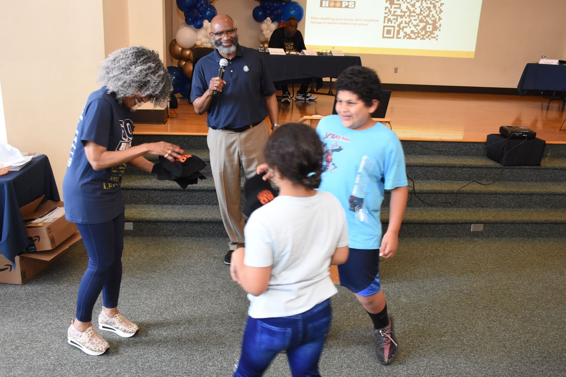 People interacting at an event: a woman hands an item to two children as a man speaks into a microphone.