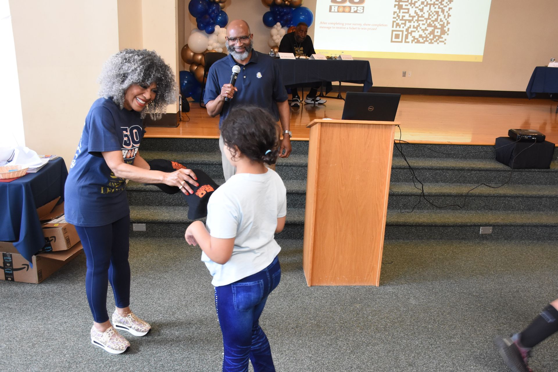 Woman hands a gift to a child at an event; a man speaks at a podium; blue and gold decorations.