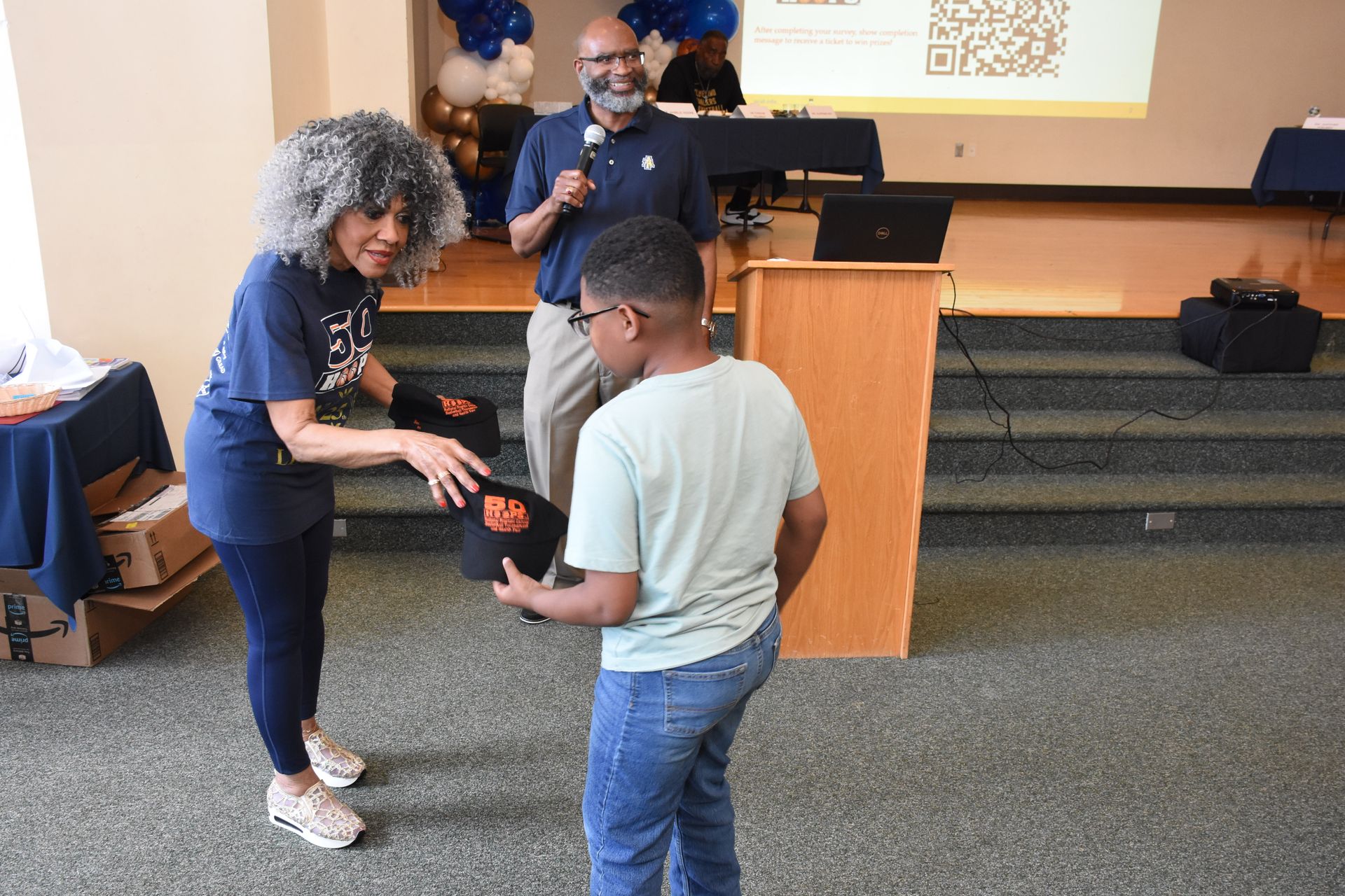 Woman giving a bag to a person while a man speaks at a podium. Event with balloons.