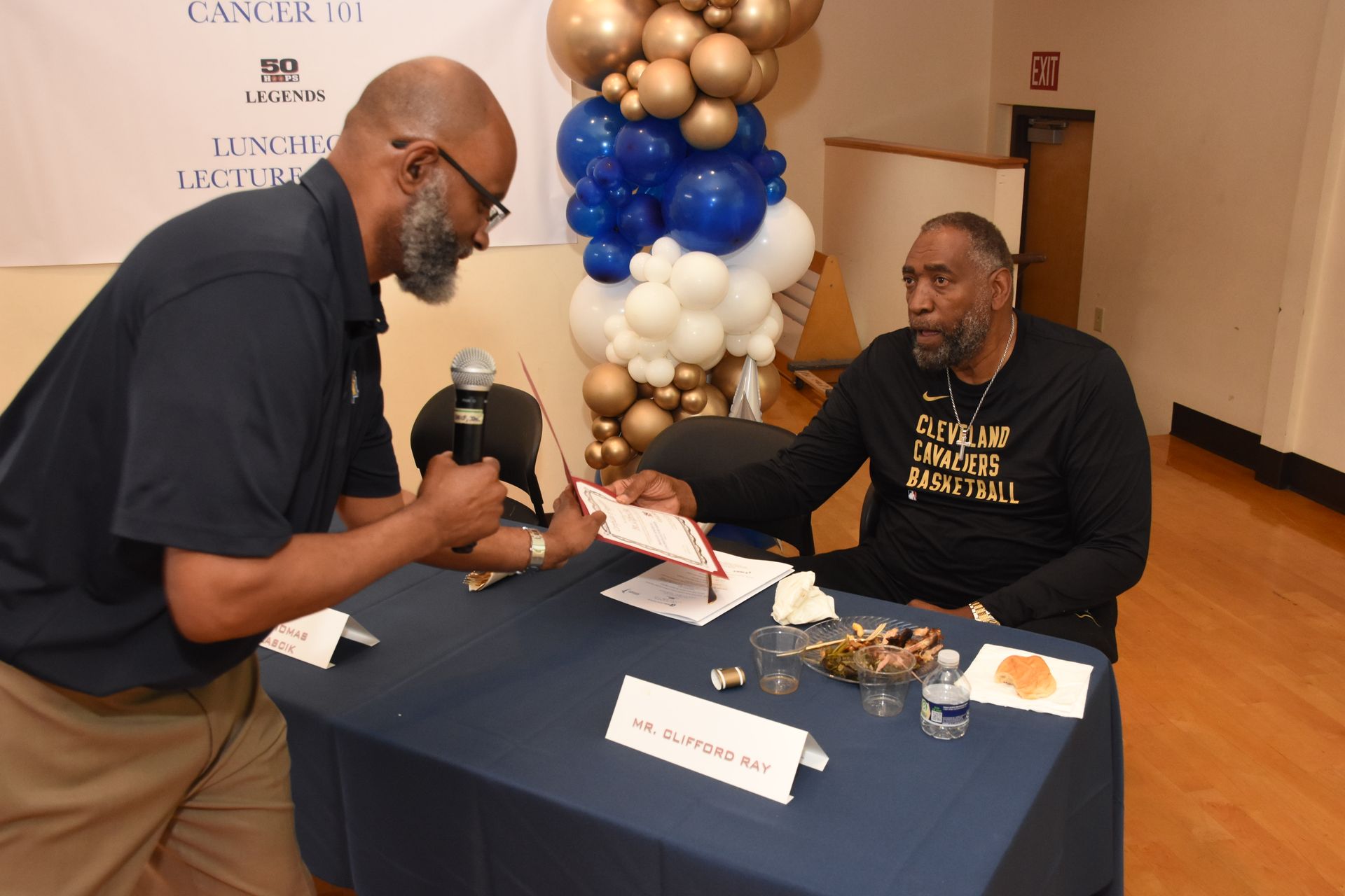 Man hands document to another man sitting at a table with balloons, a microphone, and snacks.